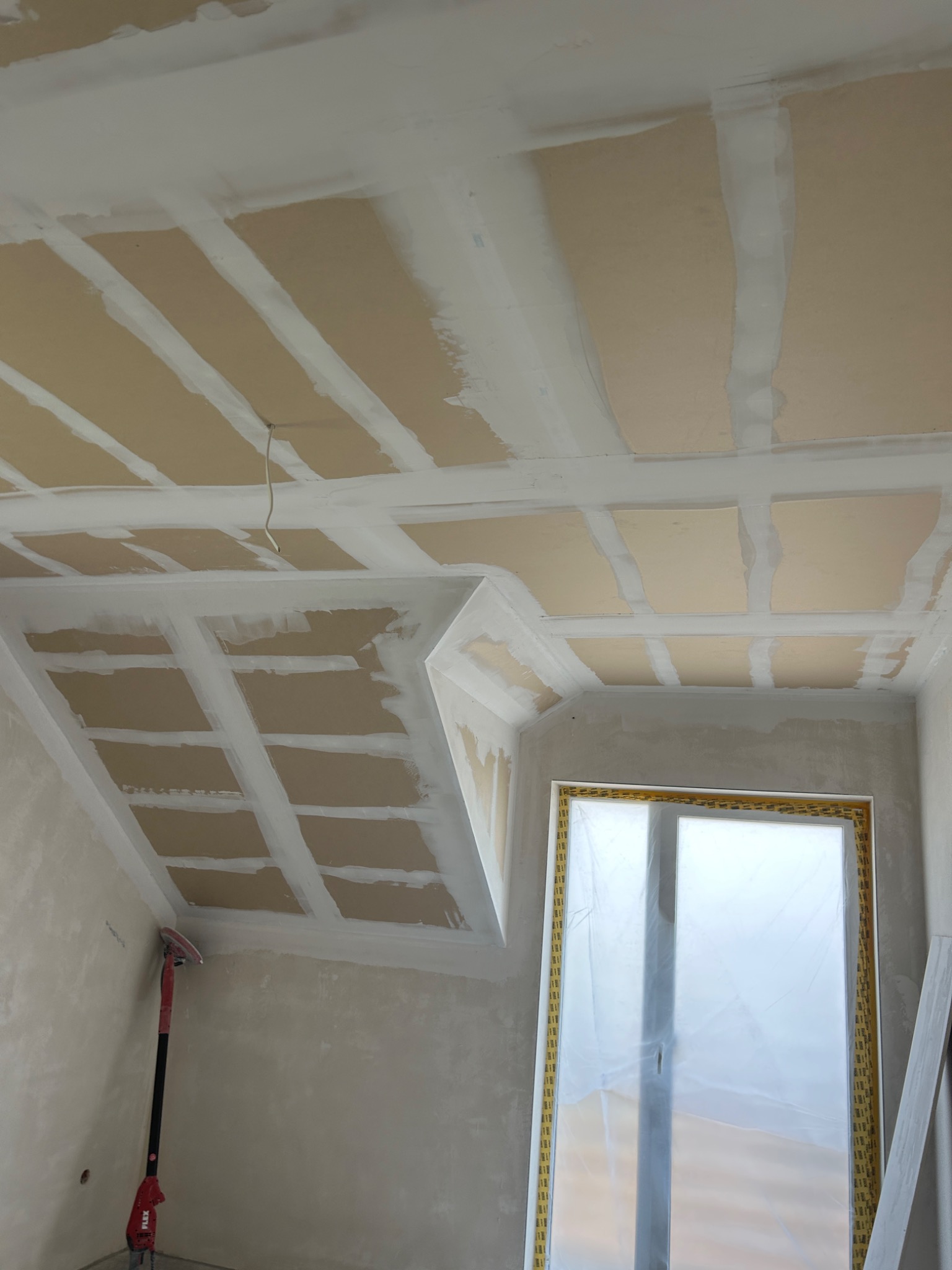 Skośny sufit z płyt gipsowych po szpachlowaniu łączeń, widoczna szlifierka do gipsu przy ścianie, okno zabezpieczone folią malarską.