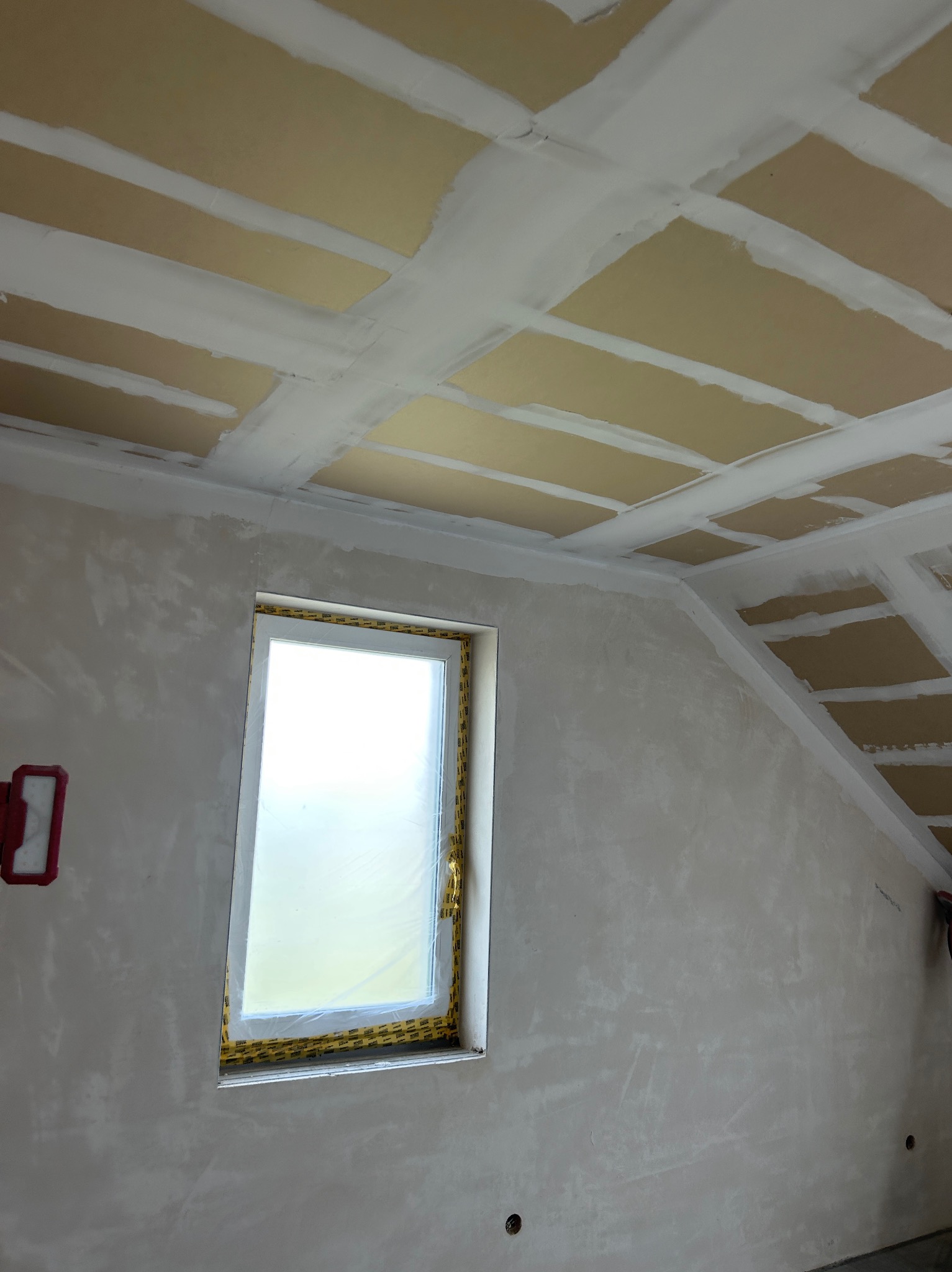 Pokój w trakcie prac wykończeniowych: ściany i sufit pokryte gładzią szpachlową, okno zabezpieczone folią. Widoczne łączenia płyt kartonowo-gipsowych.
