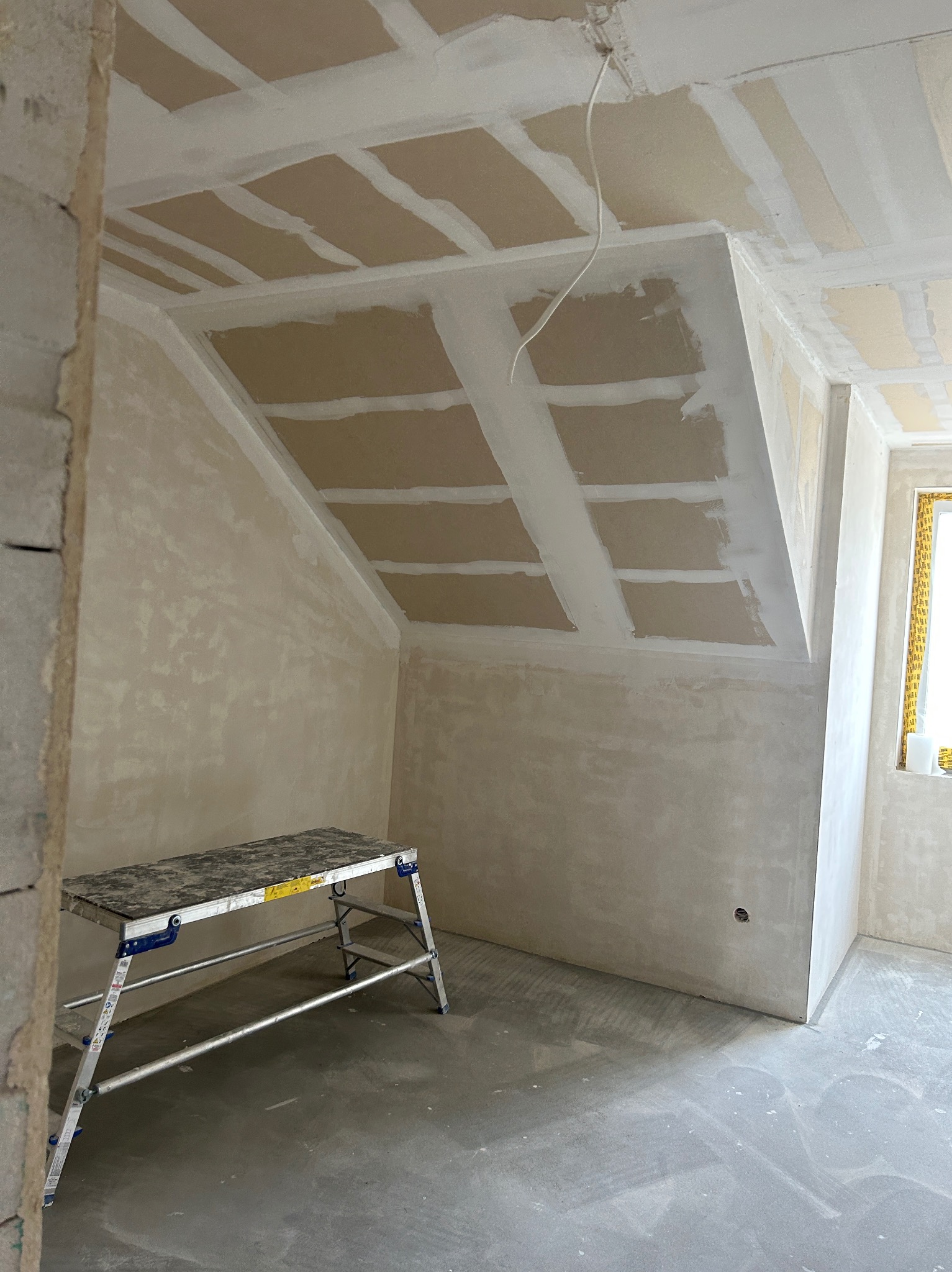 Pokój na poddaszu w trakcie prac wykończeniowych. Widoczne ściany i sufit po nałożeniu gładzi, częściowo szpachlowane płyty gipsowe. Aluminiowy podest roboczy.