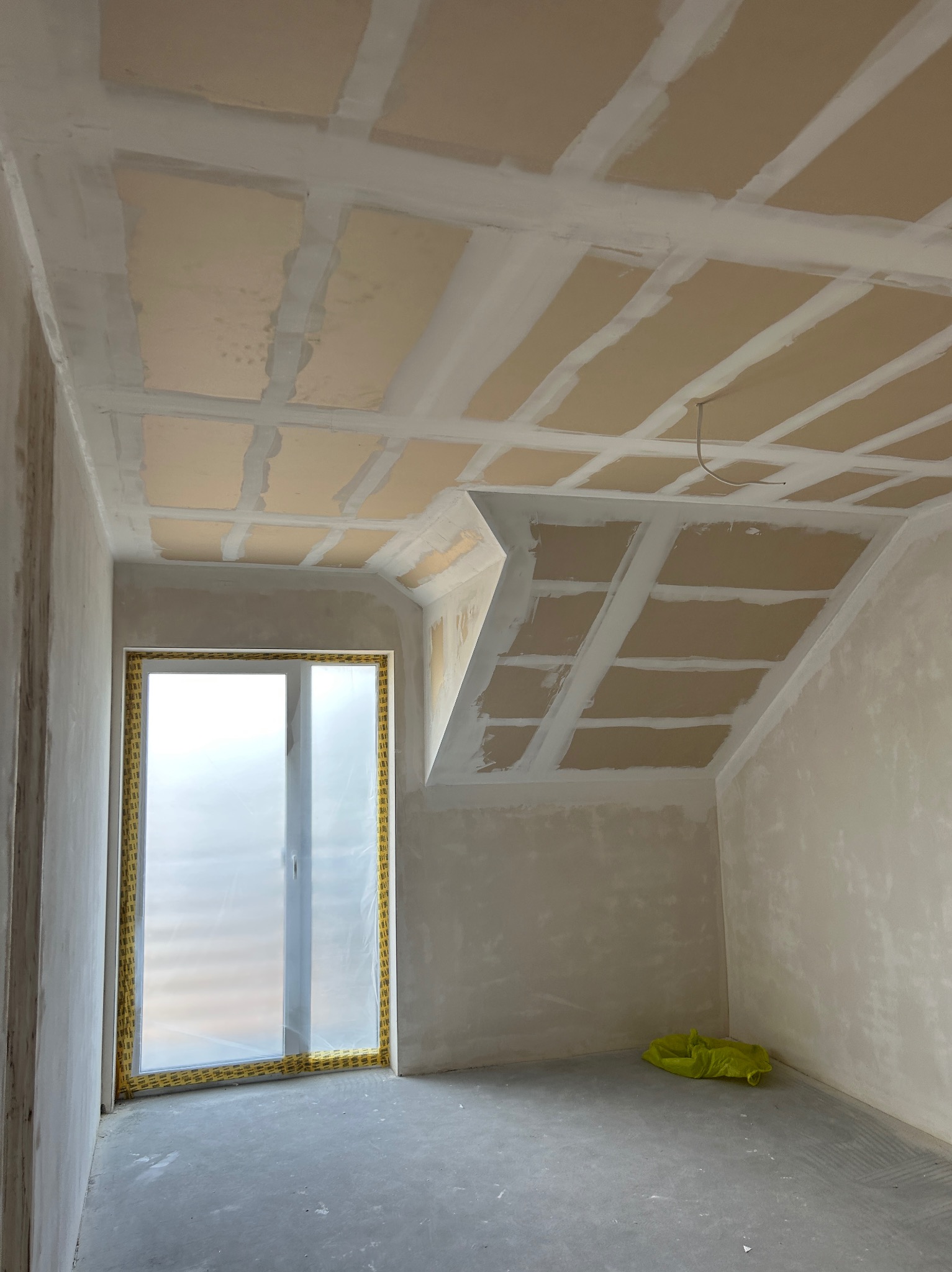 Poddasze po wykonaniu gładzi na suficie i ścianach, okno zabezpieczone folią, widoczny worek na podłodze. Stan surowy przed malowaniem.