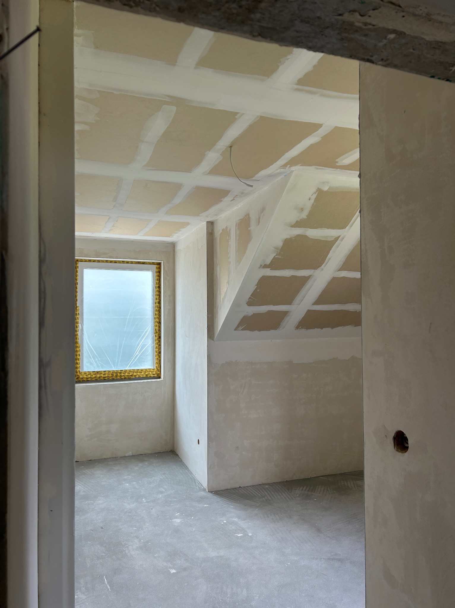 Pokój po szpachlowaniu ścian i sufitu, widok przez futrynę. Okno z folią ochronną. Przygotowanie do dalszych prac wykończeniowych.