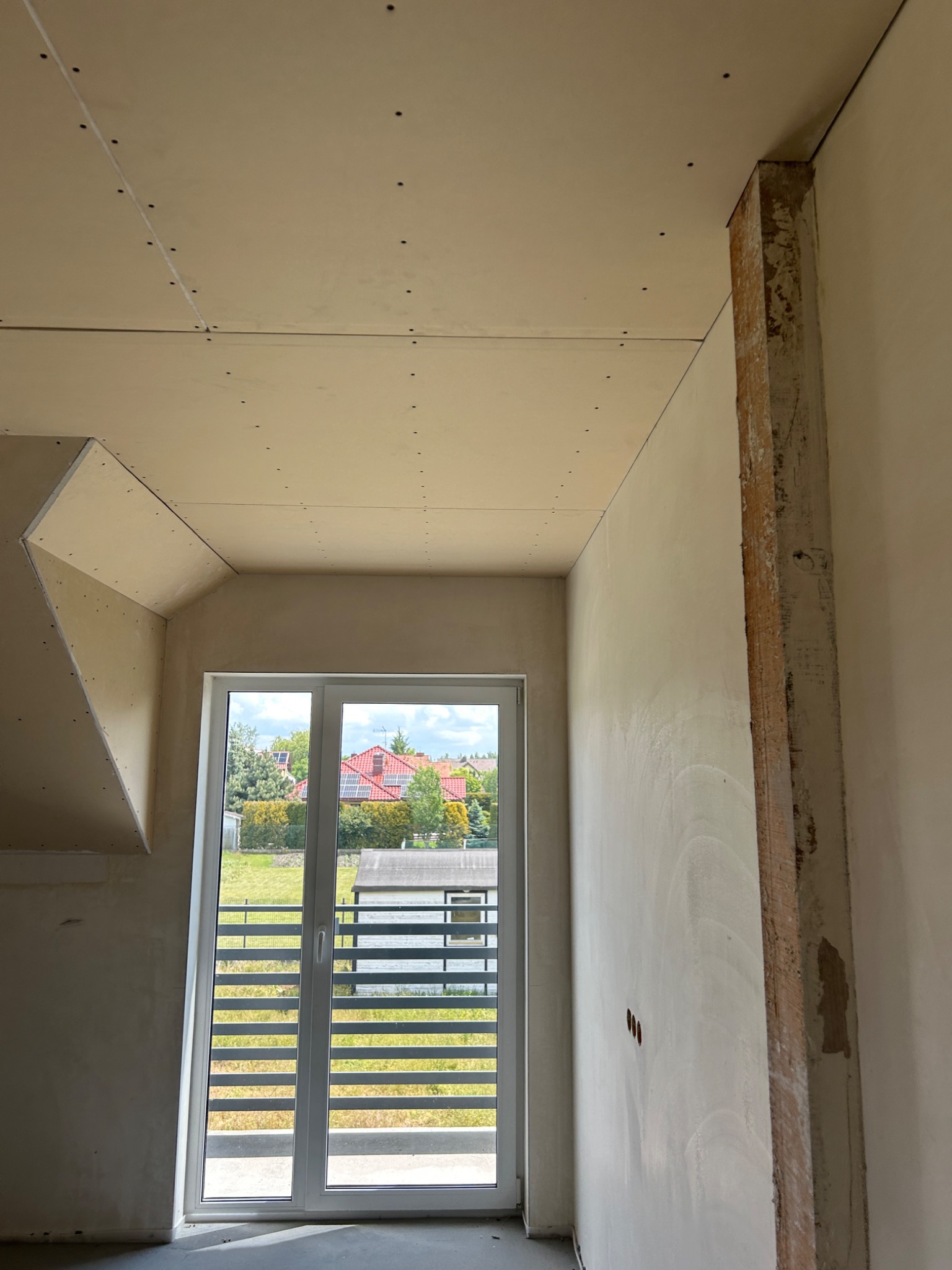 Wnętrze w trakcie wykańczania: zamontowane płyty gipsowe na suficie, ściany przygotowane pod gładź, widoczne puszki elektryczne i okno z widokiem na zewnątrz.