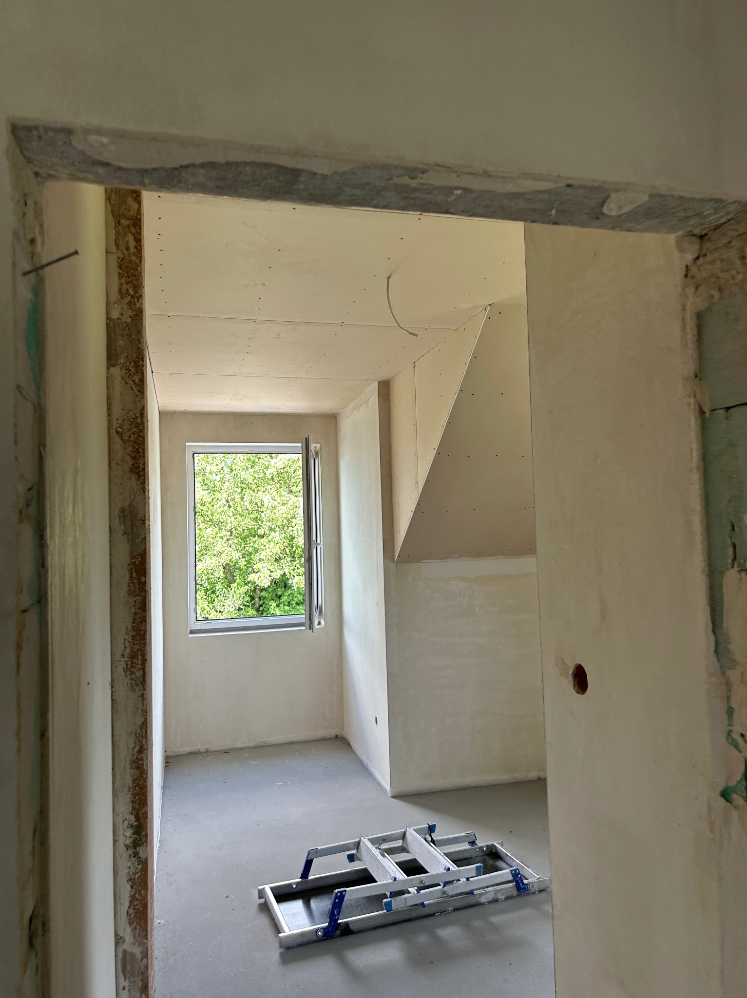 Pokój w trakcie remontu: ściany z płyt gipsowo-kartonowych, widoczne łączenia, drabina aluminiowa, otwarte okno z widokiem na zieleń.