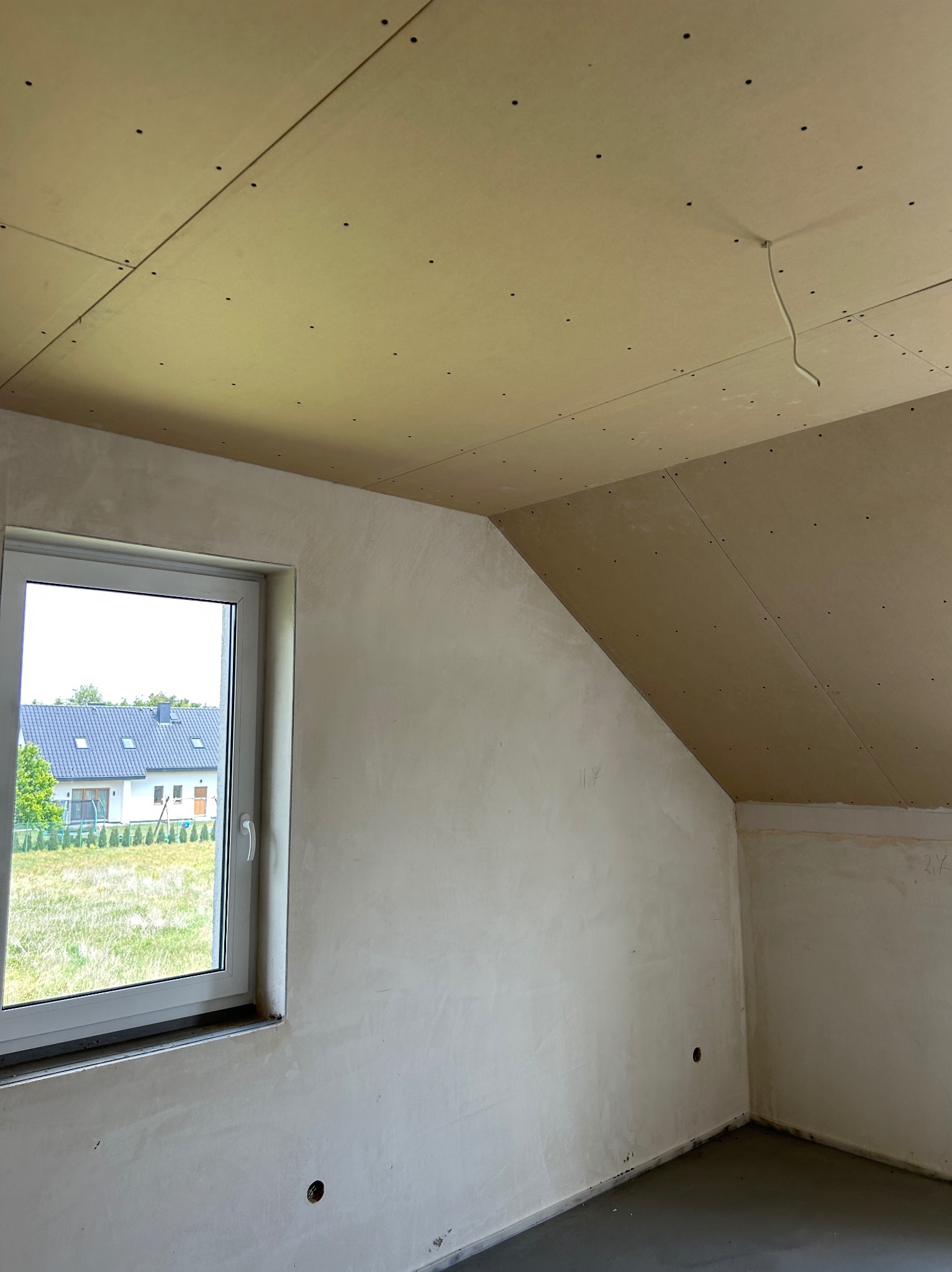 Pokój w trakcie prac wykończeniowych: ściany po nałożeniu gładzi, sufit z płyt gipsowych, okno z widokiem na dom i zieleń. Widoczne niedoskonałości i etapy prac.