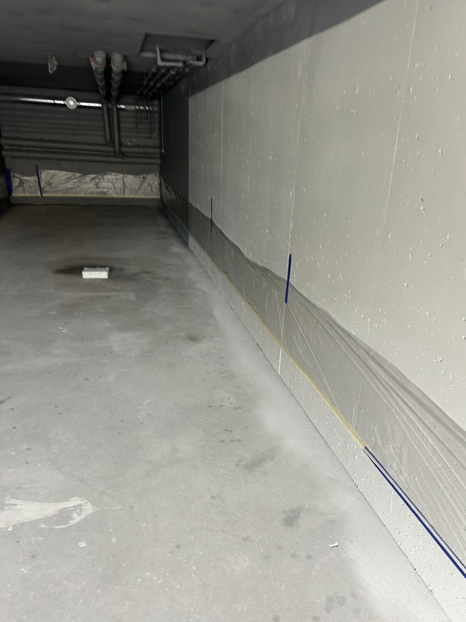 Przygotowane do malowania ściany w szarym pomieszczeniu, zabezpieczone folią i taśmą malarską. Widoczne instalacje pod sufitem.