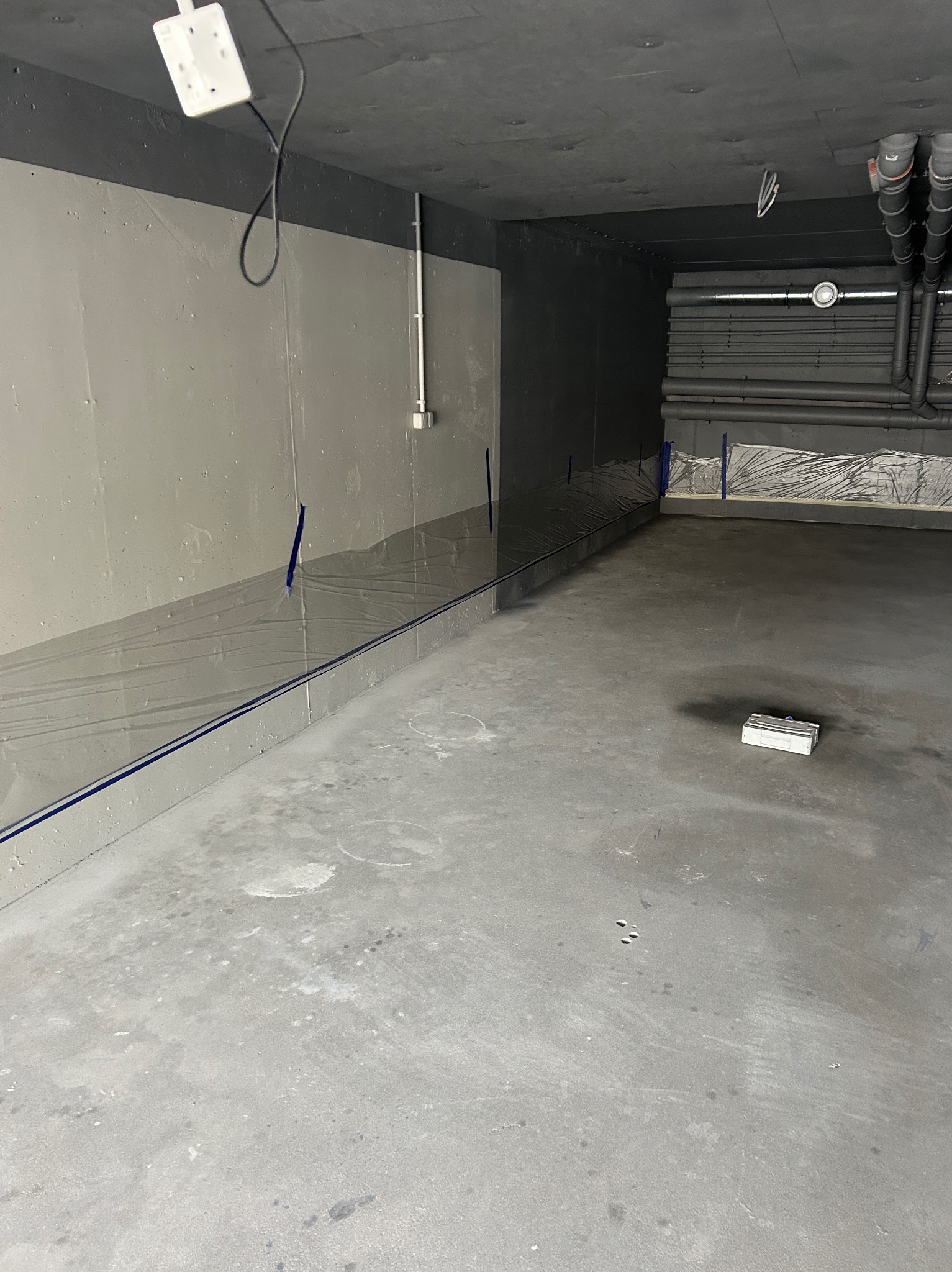 Przygotowane pomieszczenie do malowania: ściany zabezpieczone folią, widoczne rury pod sufitem, szpachlowana podłoga i narzędzia malarskie.