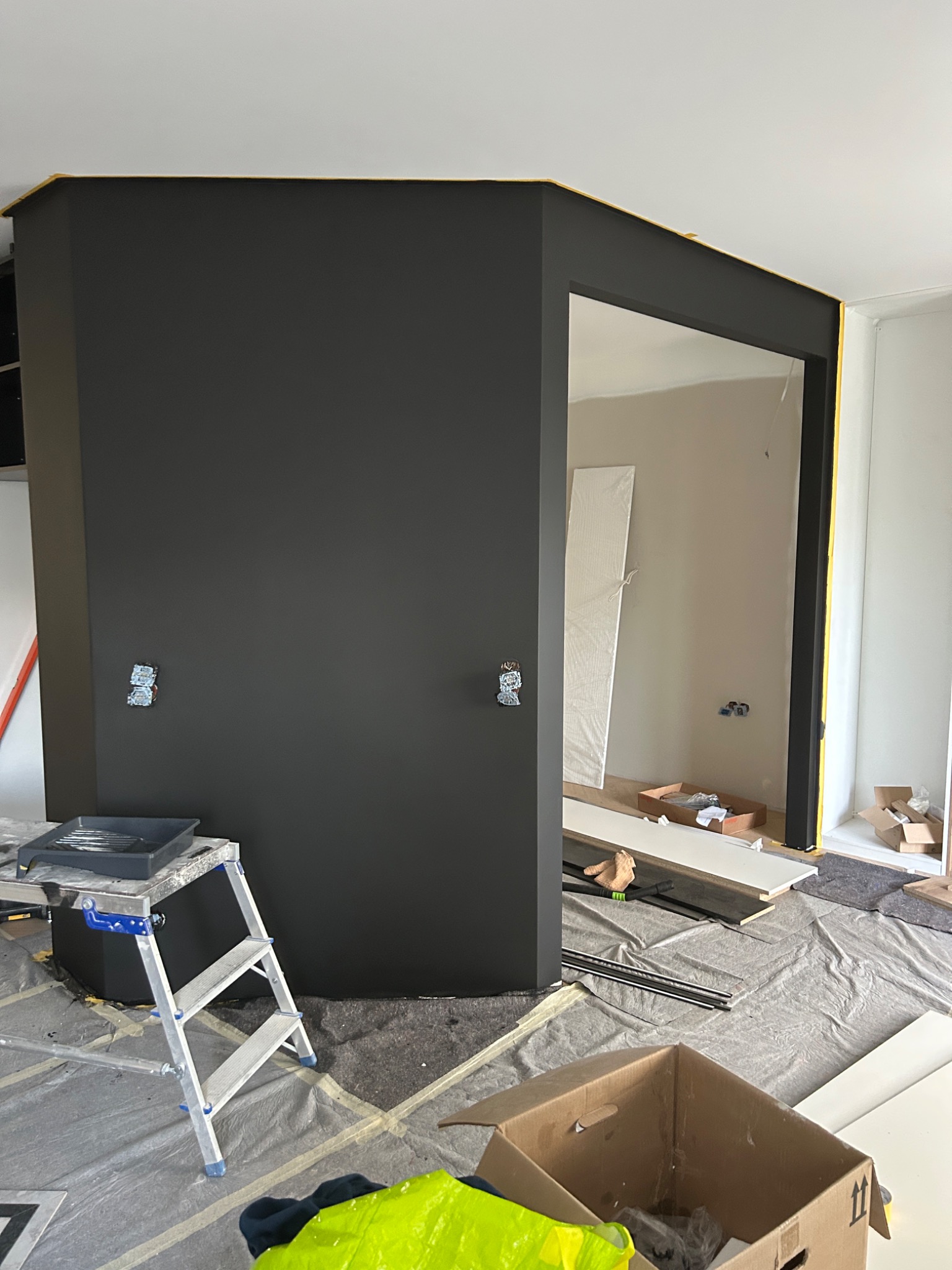 Ciemnoszara ściana w trakcie malowania z widoczną drabiną, kuwetą malarską i zabezpieczoną folią podłogą. Żółta taśma malarska na krawędziach. Świeżo pomalowane.