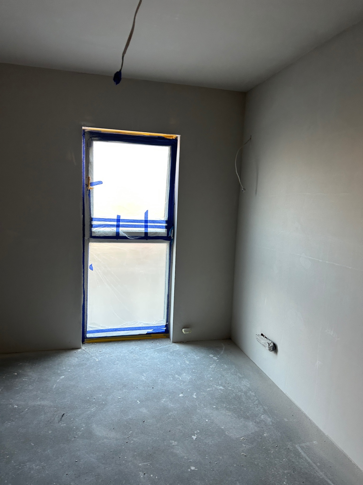 Pokój po gładziowaniu ścian, okno zabezpieczone folią i taśmą malarską. Ściany w jasnym odcieniu szarości, surowa podłoga. Widoczne przewody.