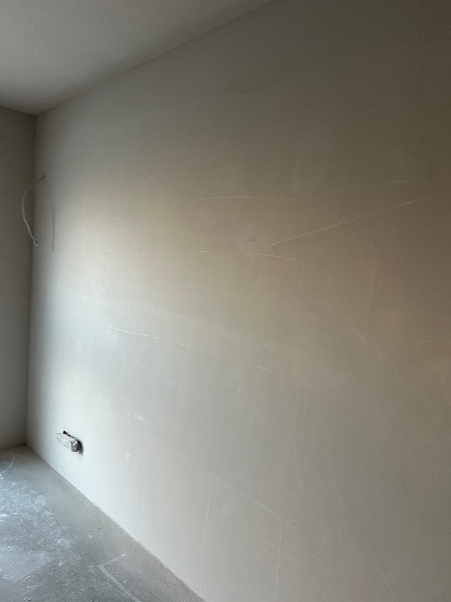 Ściana wewnętrzna po nałożeniu gładzi, widoczne ślady szpachlowania i niedoskonałości, przygotowana do malowania. Szary beton na podłodze, wystający kabel.