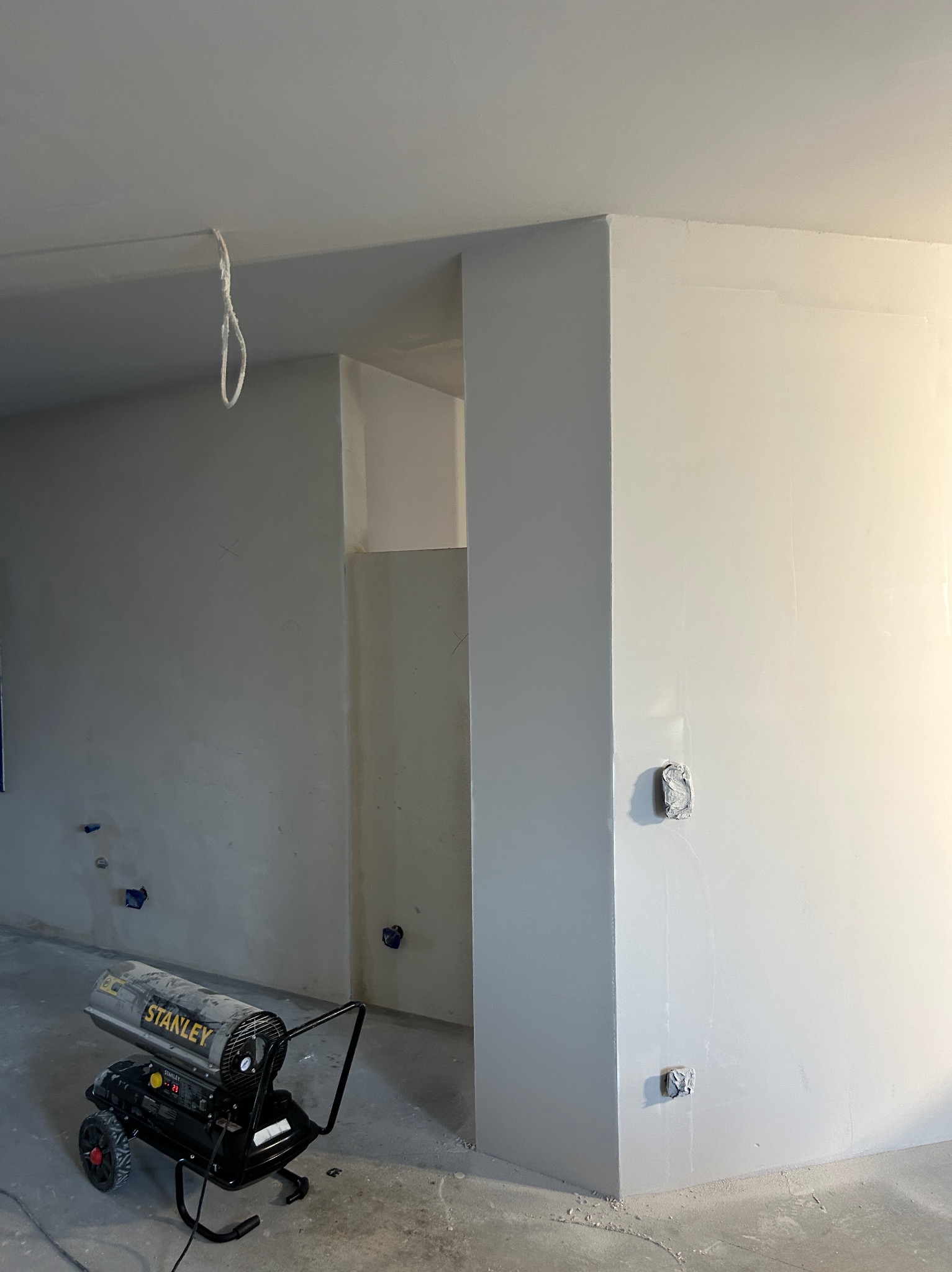 Świeżo zagruntowane ściany w pomieszczeniu z widocznym nagrzewnicą Stanley, przygotowane do dalszych prac wykończeniowych. Widoczne gniazdka zabezpieczone folią.