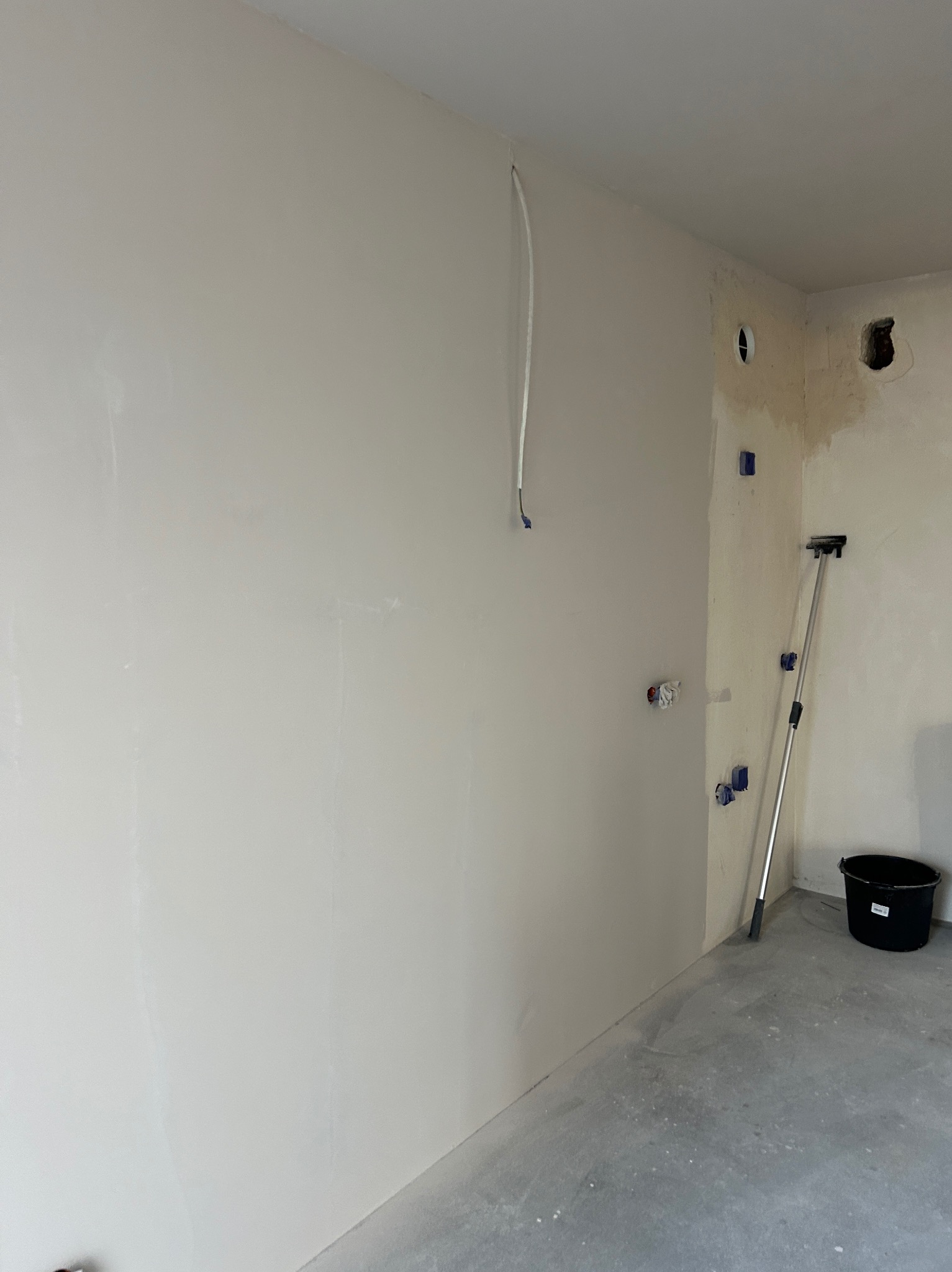 Wnętrze po gładziowaniu ścian, widoczne zabezpieczone instalacje, przygotowane do dalszych prac wykończeniowych. Na podłodze stoi wiadro i narzędzie.