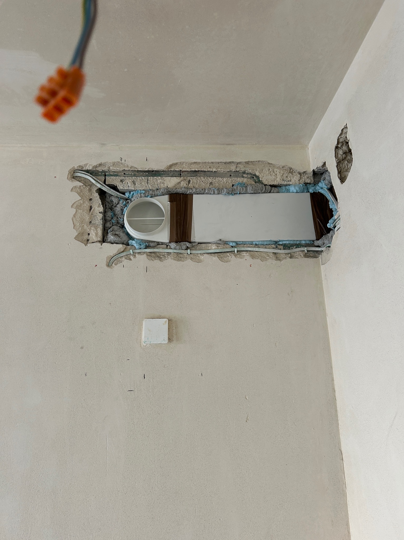Biała ściana z otworem na wentylację, zabezpieczona pianką montażową i kable. Widoczny fragment instalacji elektrycznej i gniazdko. Prace remontowe w toku.