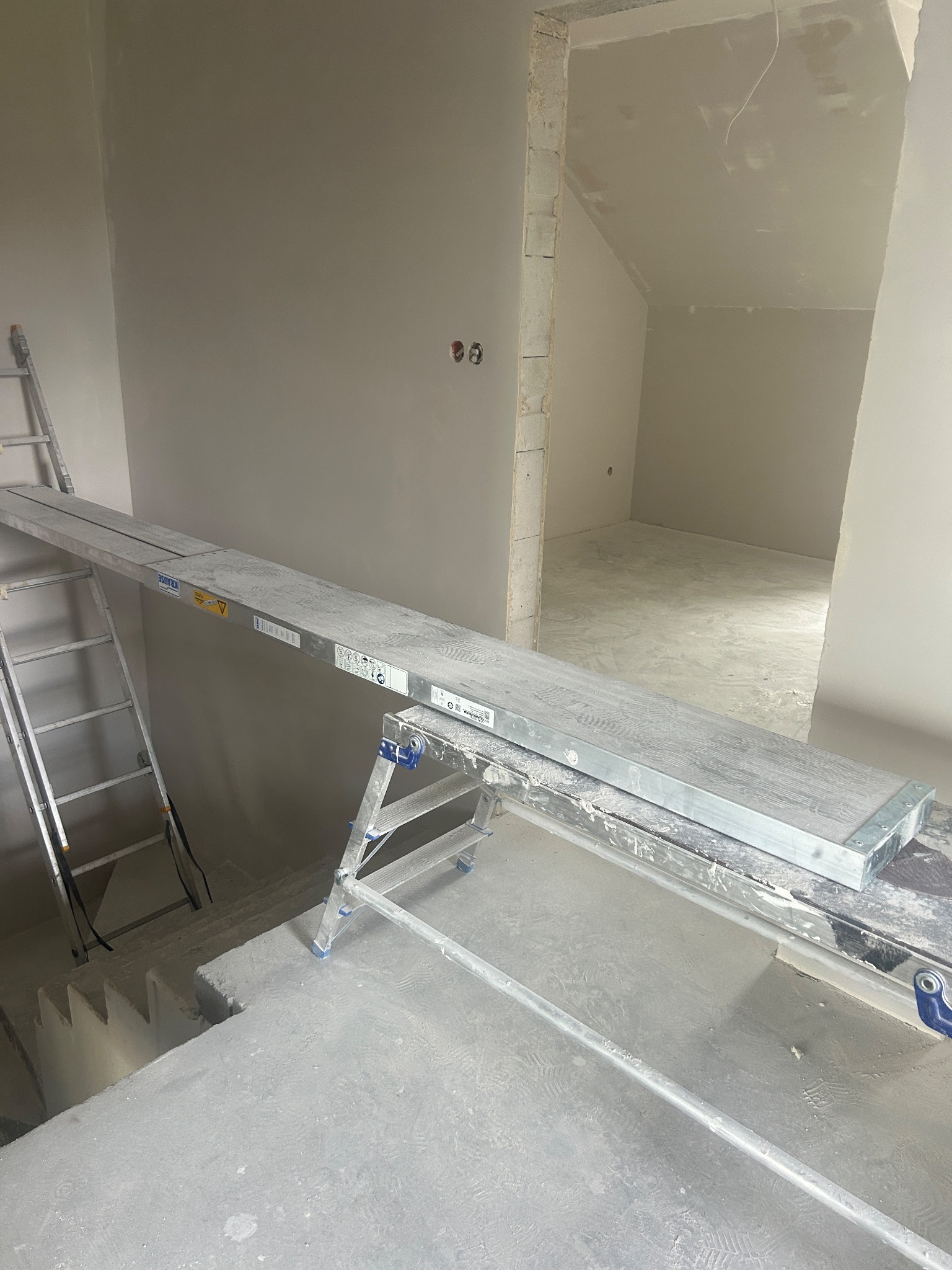 Wnętrze w trakcie prac wykończeniowych: ściany po nałożeniu gładzi, widoczne rusztowanie i drabina, przygotowane do dalszych prac malarskich. Surowy stan wykończenia.