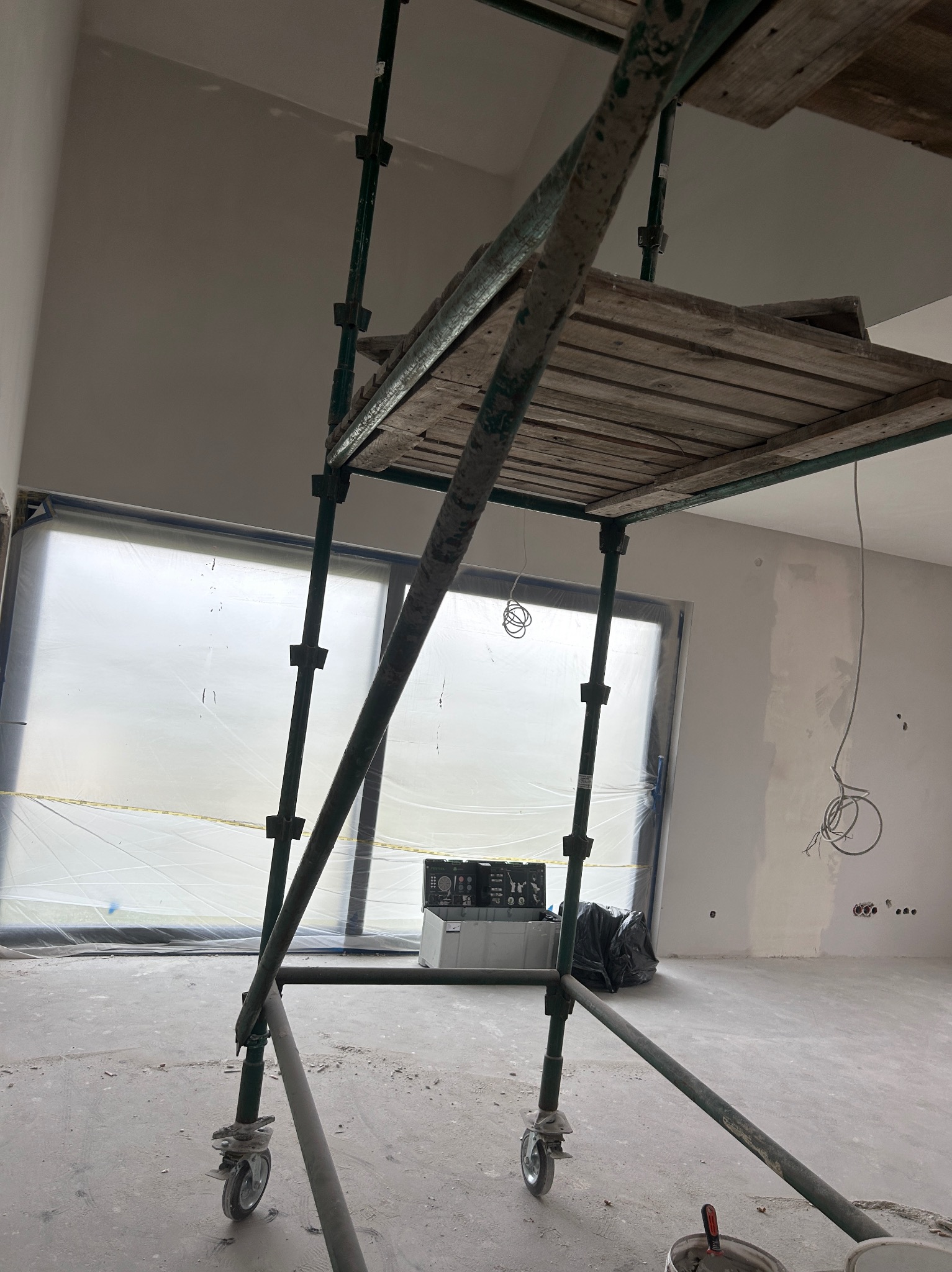 Wnętrze w trakcie remontu: rusztowanie na tle okna z folią ochronną, ściany częściowo pokryte gładzią, widoczne kable elektryczne. Etap prac wykończeniowych.