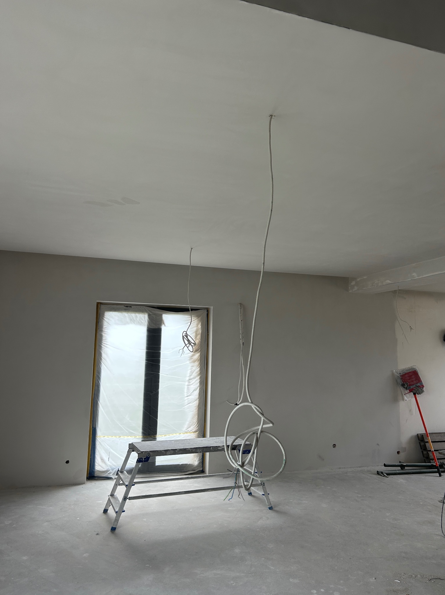 Wnętrze w trakcie prac wykończeniowych: gładkie ściany, okno zabezpieczone folią, drabina i wiszące kable elektryczne. Widoczne ślady szpachlowania na suficie.