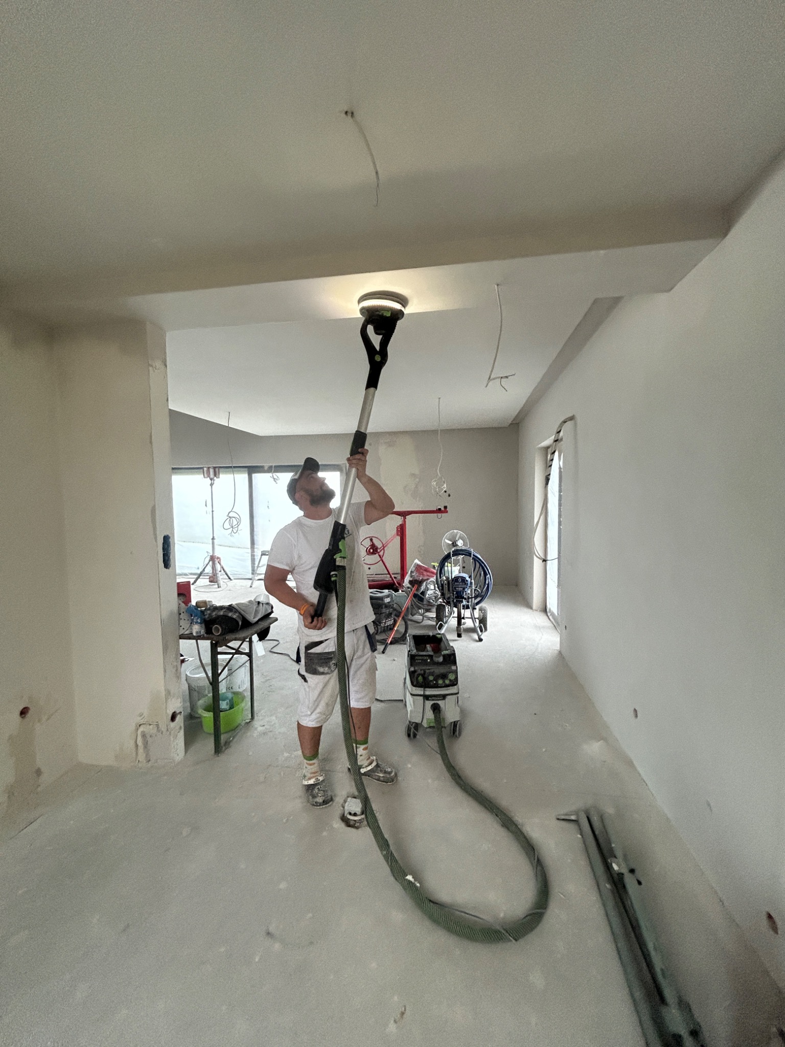 Pracownik szlifuje sufit szlifierką teleskopową w jasnym wnętrzu. Widoczne narzędzia, przewody i przygotowane ściany. Praca w toku, remont mieszkania.