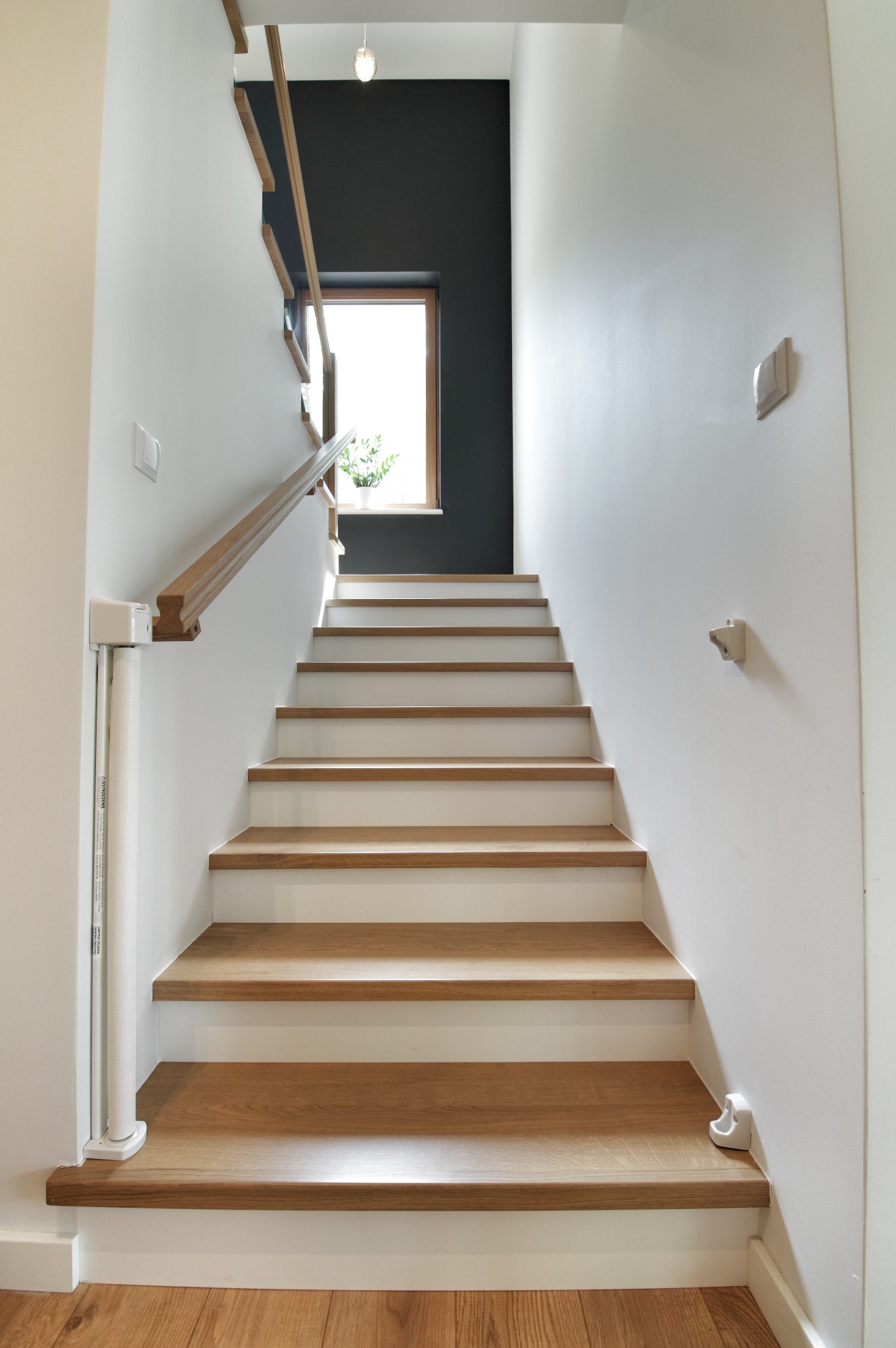 Nowoczesne schody z drewnianymi stopniami i białymi podstopnicami prowadzące do okna z widokiem na zieleń, zabezpieczenie dla dzieci zamontowane przy poręczy.