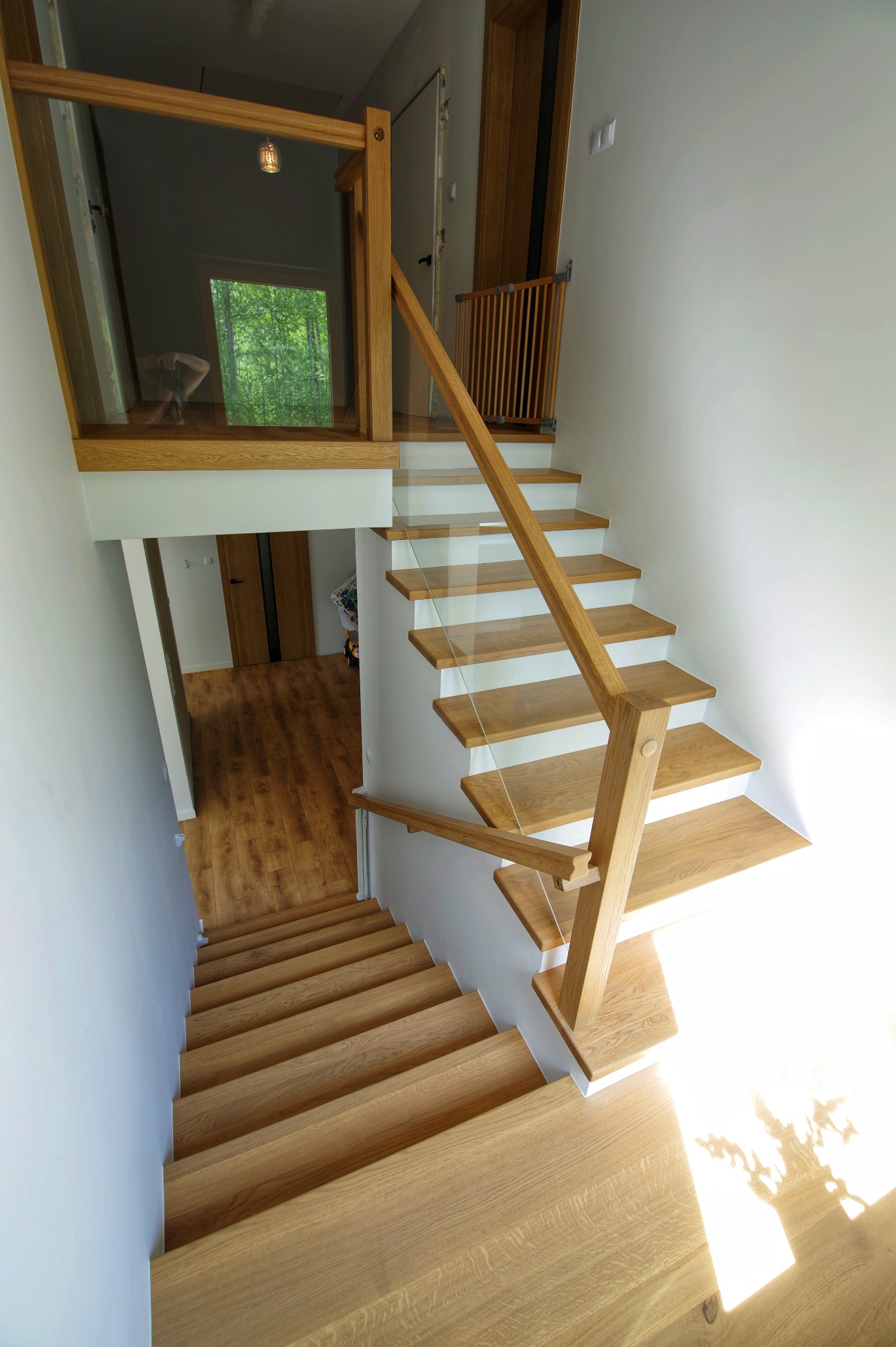 Nowoczesne schody drewniane z jasnego drewna z szklaną balustradą i drewnianą poręczą, widok z góry na dół, z widoczną bramką zabezpieczającą dla dzieci na górnym podeście.