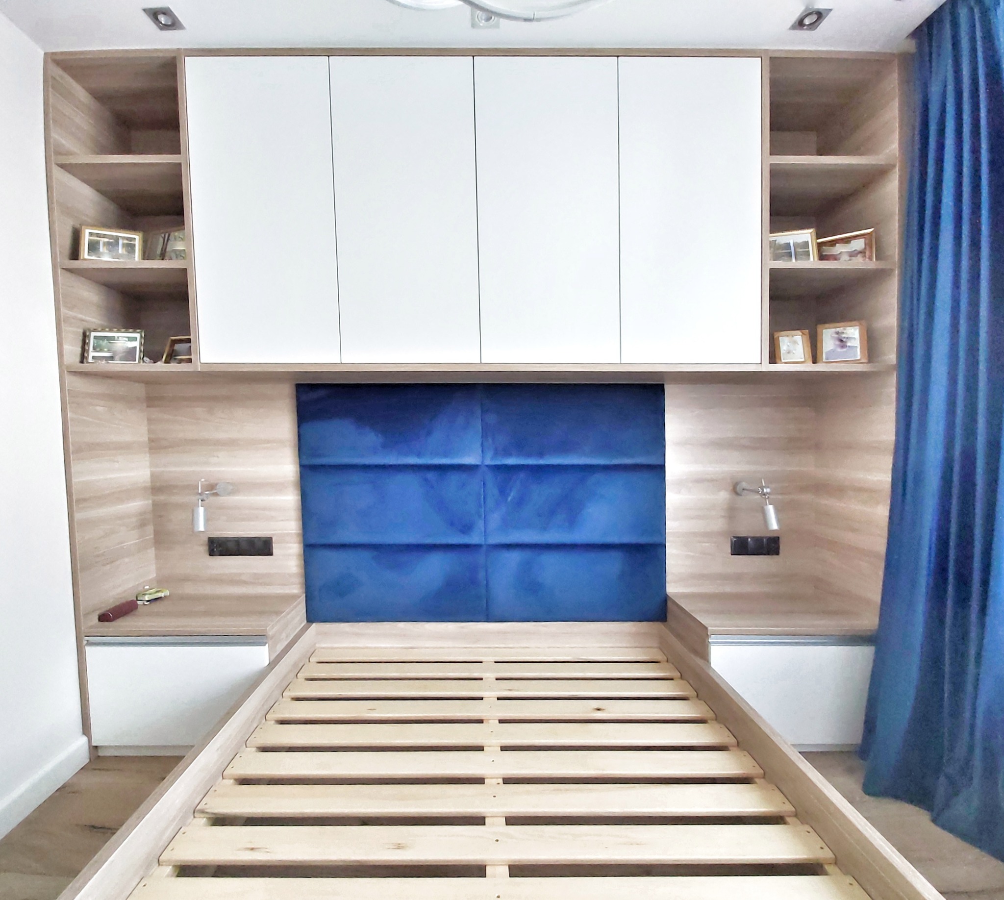 Rama łóżka z drewnianym stelażem, wbudowana w zabudowę meblową z szafkami, półkami i oświetleniem punktowym, z niebieskim panelem tapicerowanym jako wezgłowie