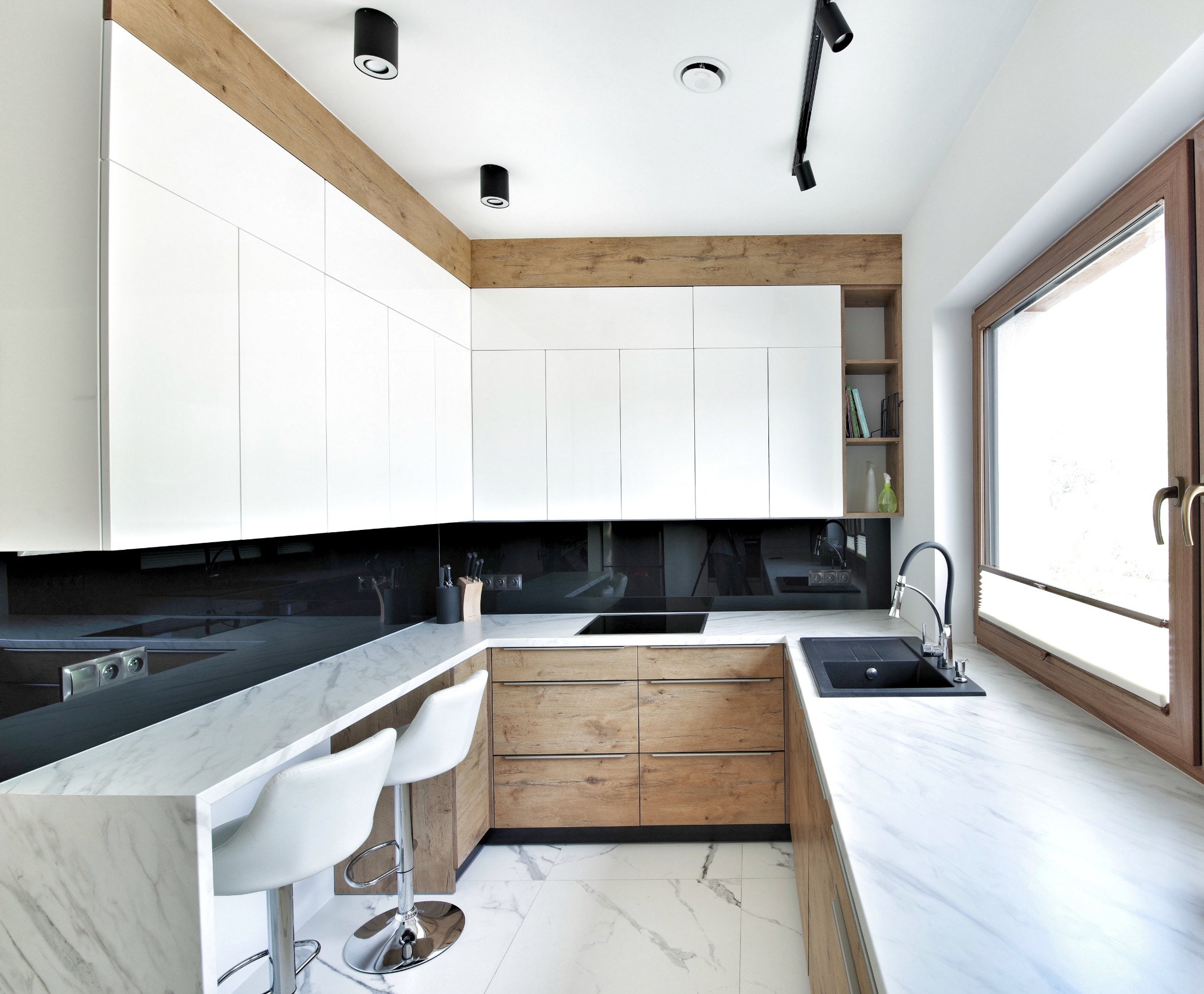Nowoczesna kuchnia z białymi szafkami górnymi, drewnianymi dolnymi, marmurowym blatem i podłogą, czarnym panelem nad blatem oraz czarnym zlewem pod oknem.