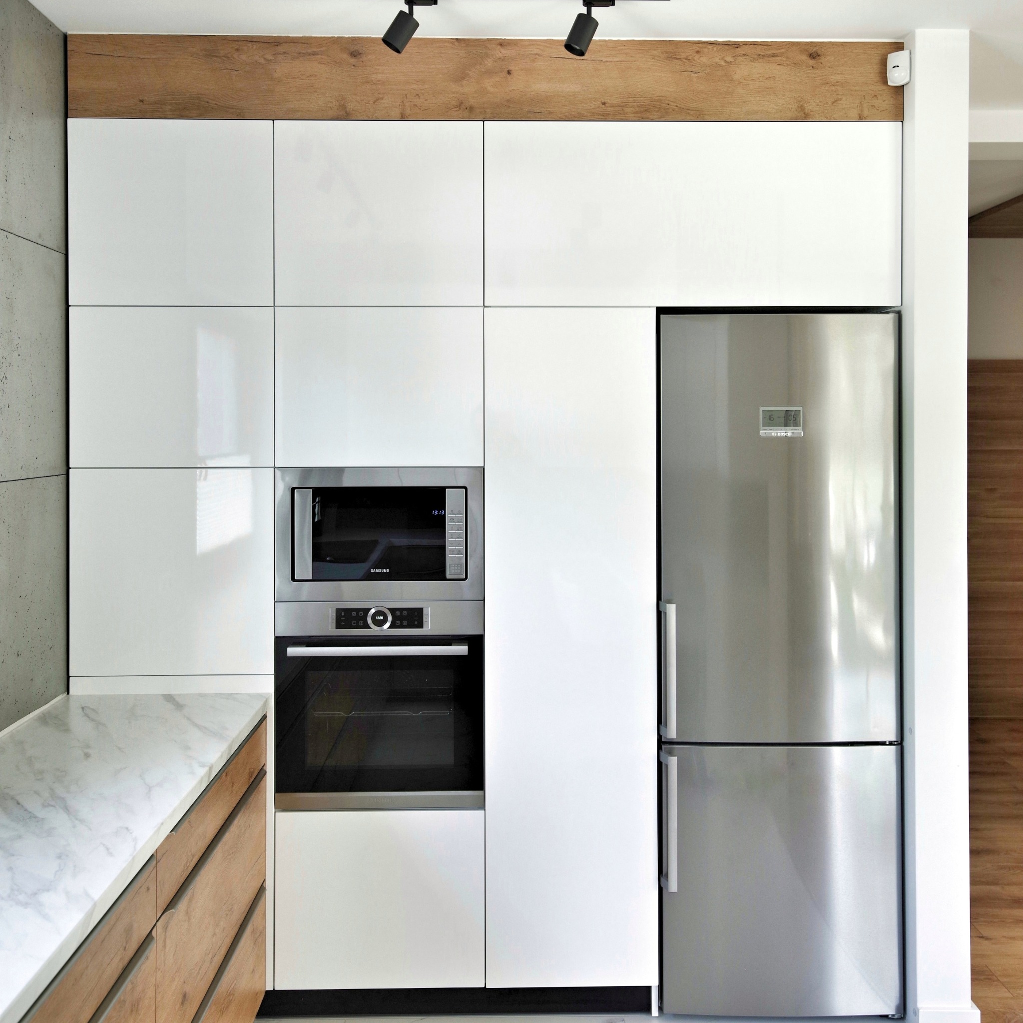 Nowoczesna kuchnia z białymi, lakierowanymi frontami, marmurowym blatem, zabudowanym sprzętem AGD (piekarnik, mikrofalówka) i dużą, dwudrzwiową lodówką ze stali nierdzewnej.