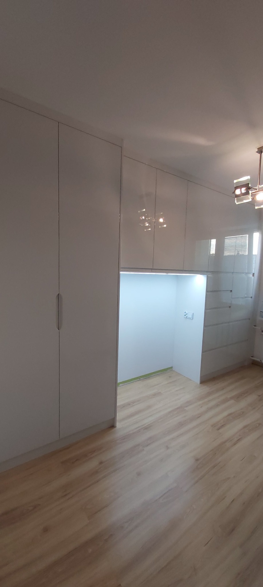 Biała zabudowa meblowa z szafami i wnęką z oświetleniem LED, widoczna drewniana podłoga i nowoczesna lampa sufitowa.