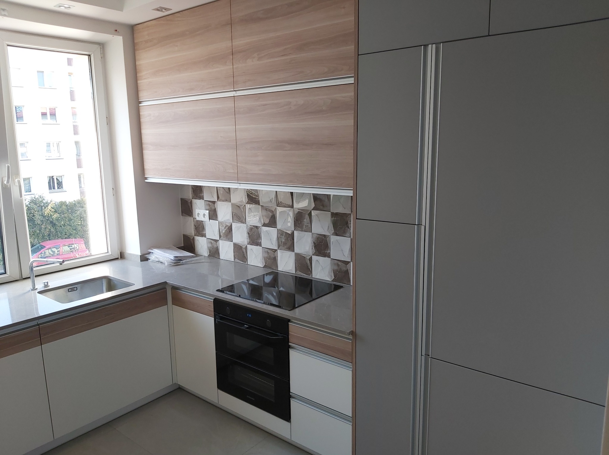 Nowoczesna kuchnia z białymi i drewnopodobnymi szafkami, szarym blatem i płytkami w geometryczny wzór na ścianie, z widokiem na budynek mieszkalny za oknem.
