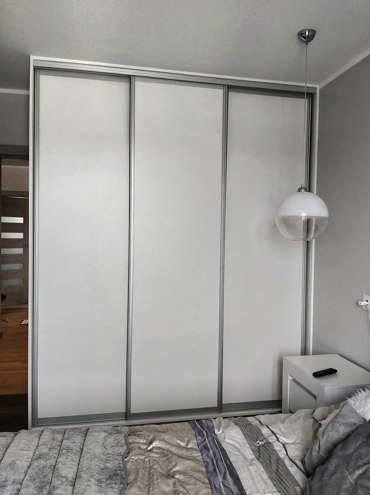 Biała szafa przesuwna z trzema drzwiami, srebrne obramowanie, widoczna szafka nocna z pilotem i lampa wisząca w sypialni.