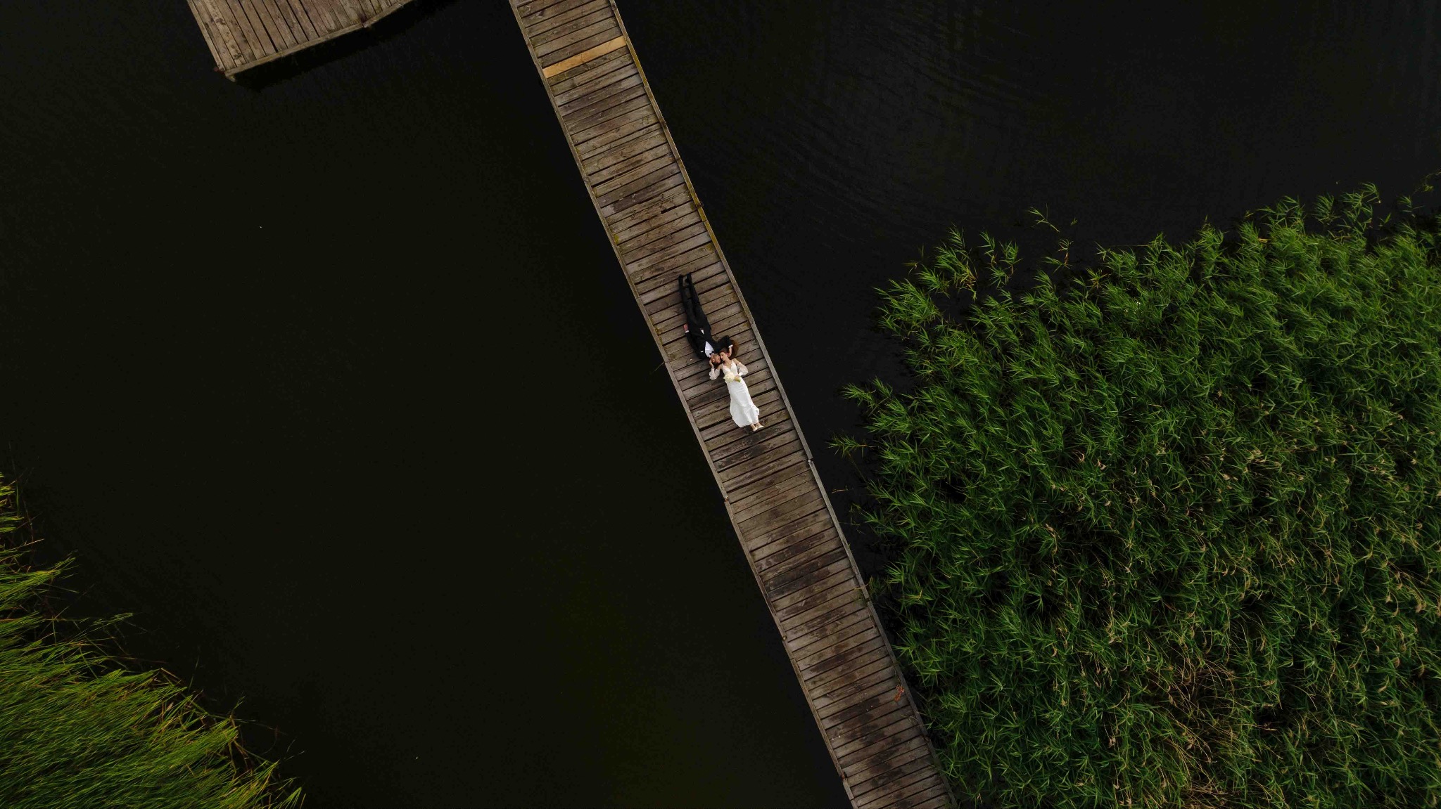 Para młoda leży na drewnianym pomoście nad wodą, widok z góry, otoczona trzcinami.