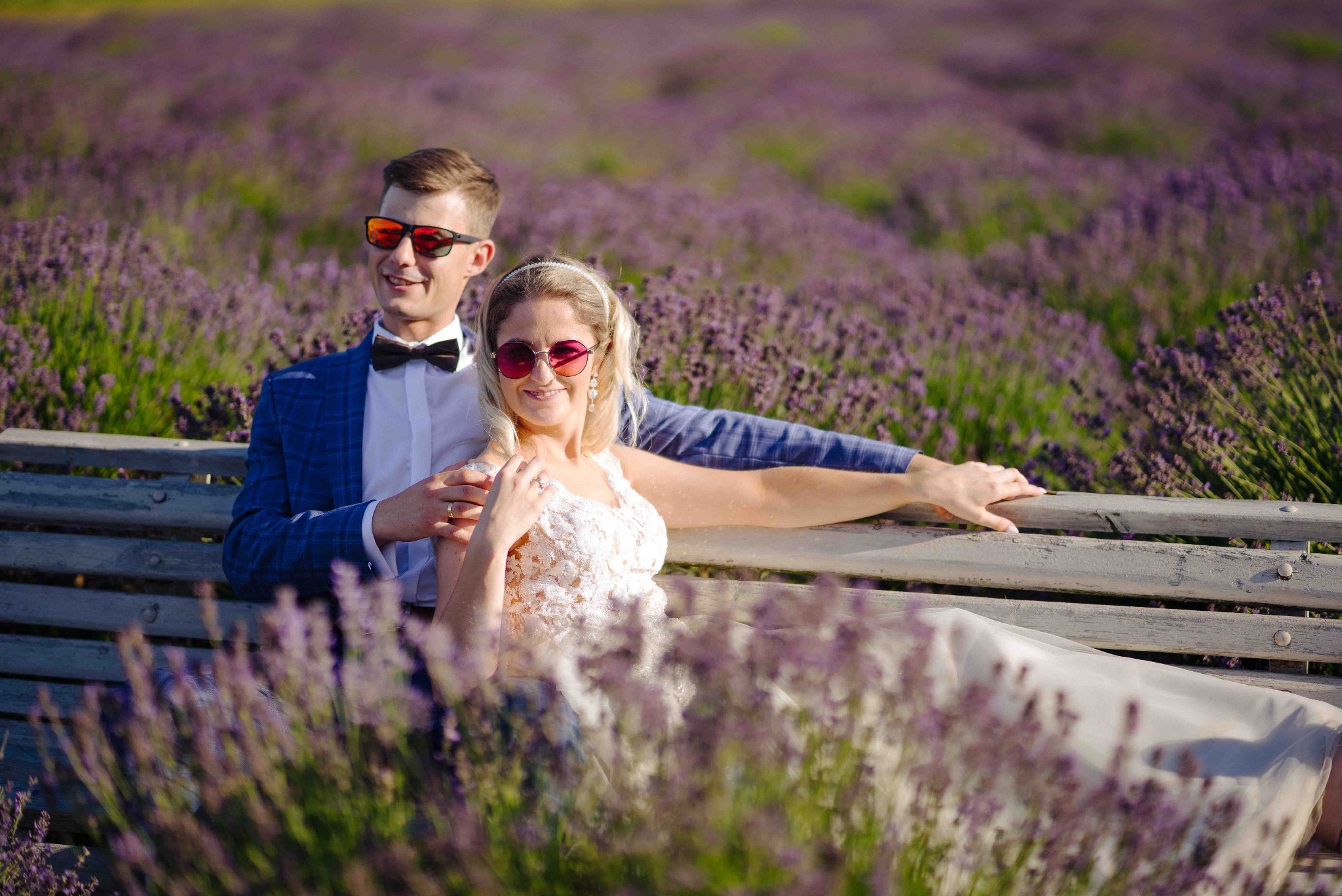 Para młoda w okularach przeciwsłonecznych siedzi na ławce w polu lawendy, mężczyzna w niebieskiej marynarce w kratę i muszce, kobieta w koronkowej sukni ślubnej.