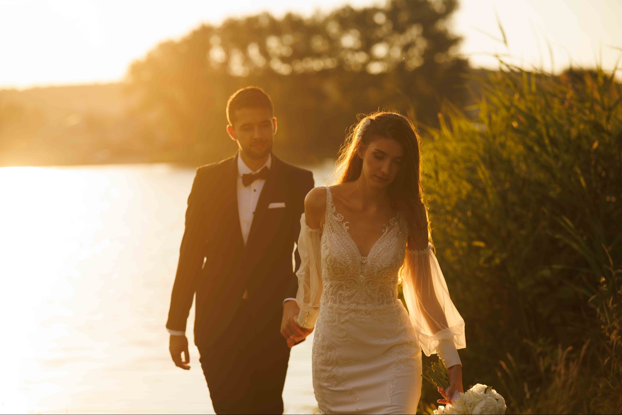 Para młoda, on w czarnym garniturze z muchą, ona w białej sukni ślubnej z bukietem, spacerują brzegiem jeziora w promieniach zachodzącego słońca.