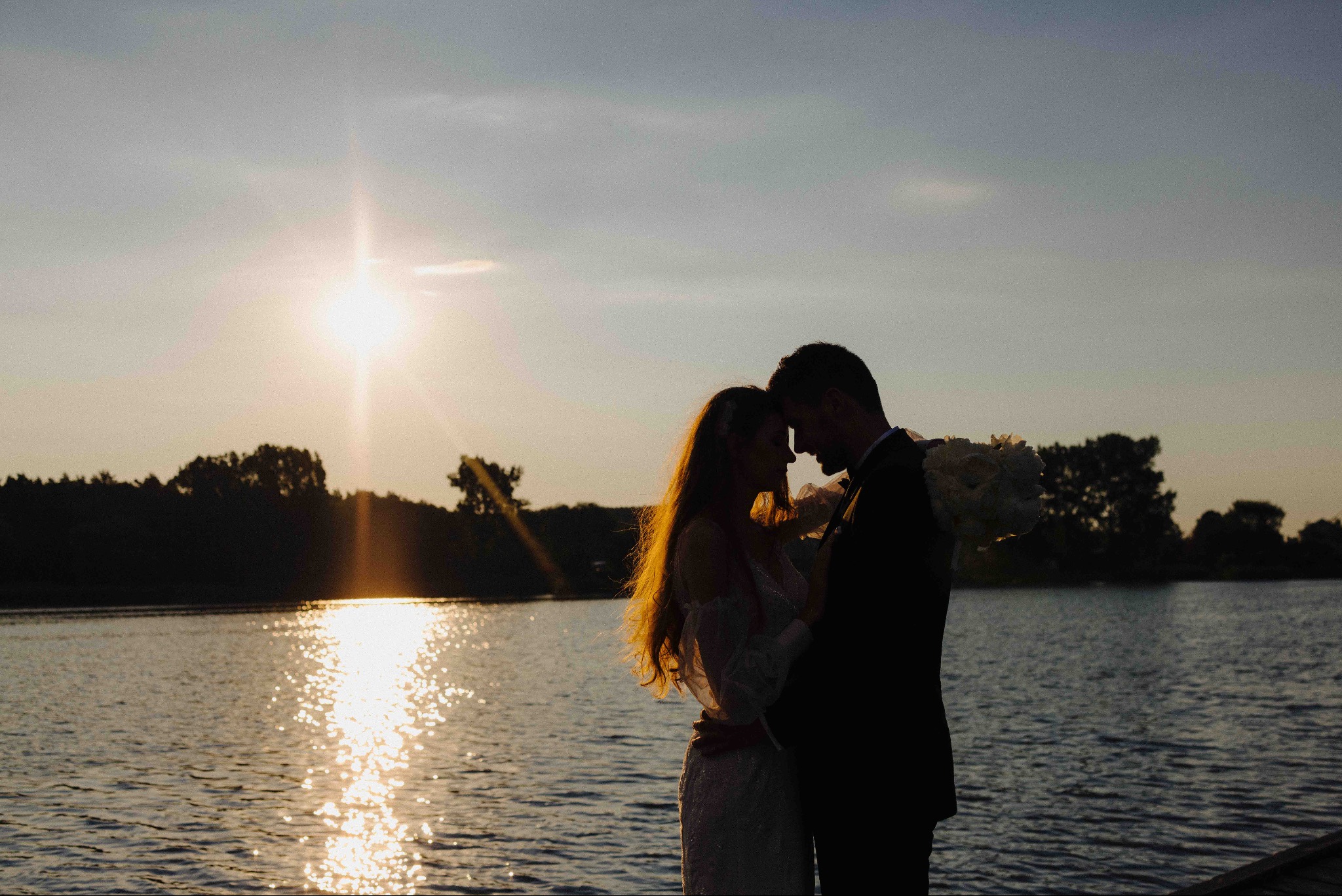 Silhouettes of a bride and groom embracing against a sunset over a lake, the sun reflecting on the water's surface, creating a romantic and intimate atmosphere.
