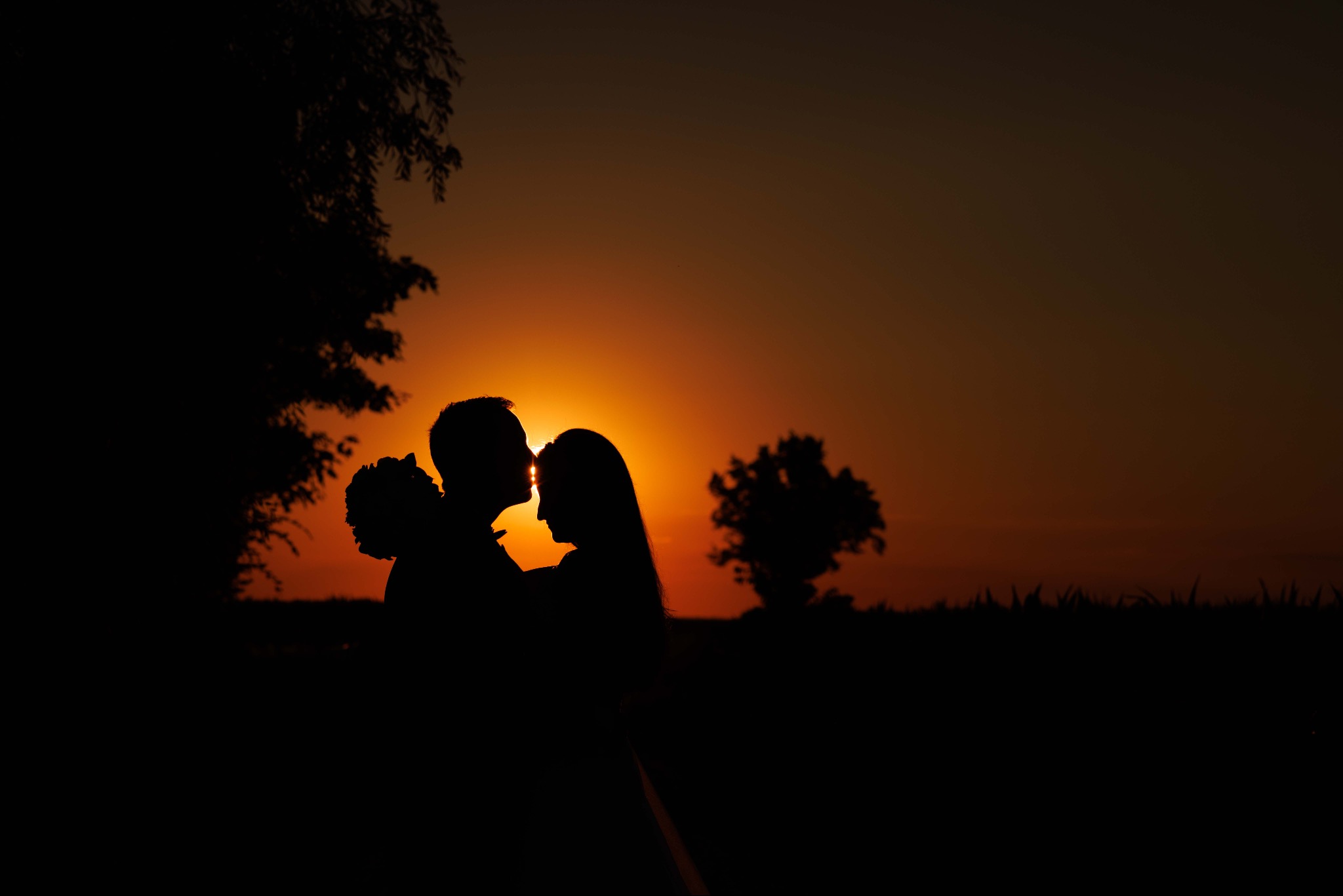 Sylwetki pary młodej podczas pocałunku na tle zachodzącego słońca w Strykowie, zarys drzew w tle tworzy romantyczny klimat.