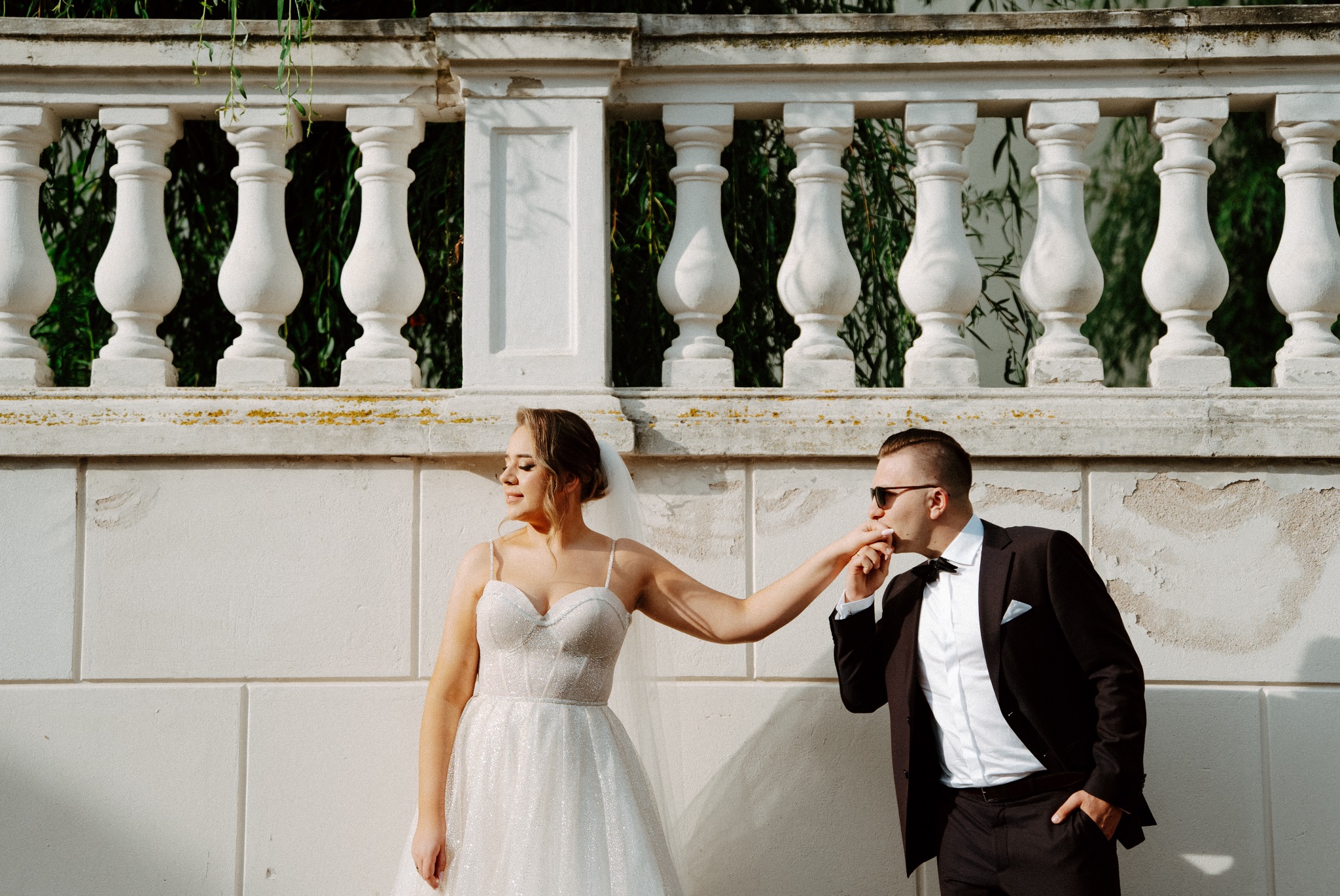 Panna młoda w białej sukni ślubnej z brokatem, stojąca na tle białej balustrady z zielenią w tle, pan młody w czarnym garniturze całuje jej dłoń, oboje w promieniach słońca.