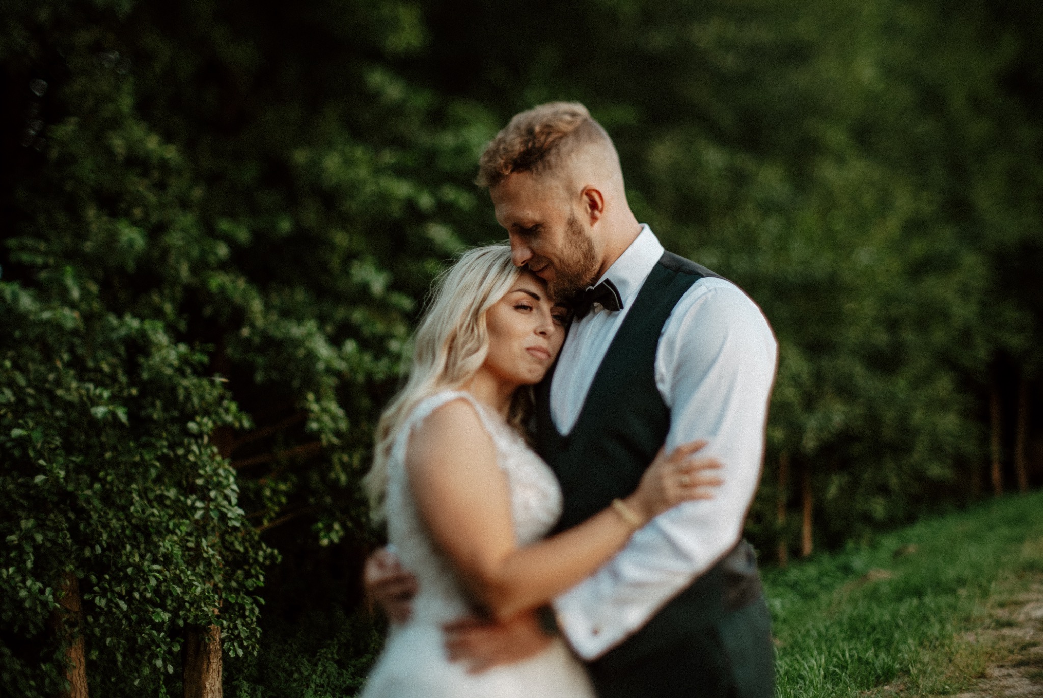 Para młoda w objęciach na tle zieleni, panna młoda w sukni z koronkowymi zdobieniami, pan młody w czarnej muszce i kamizelce.