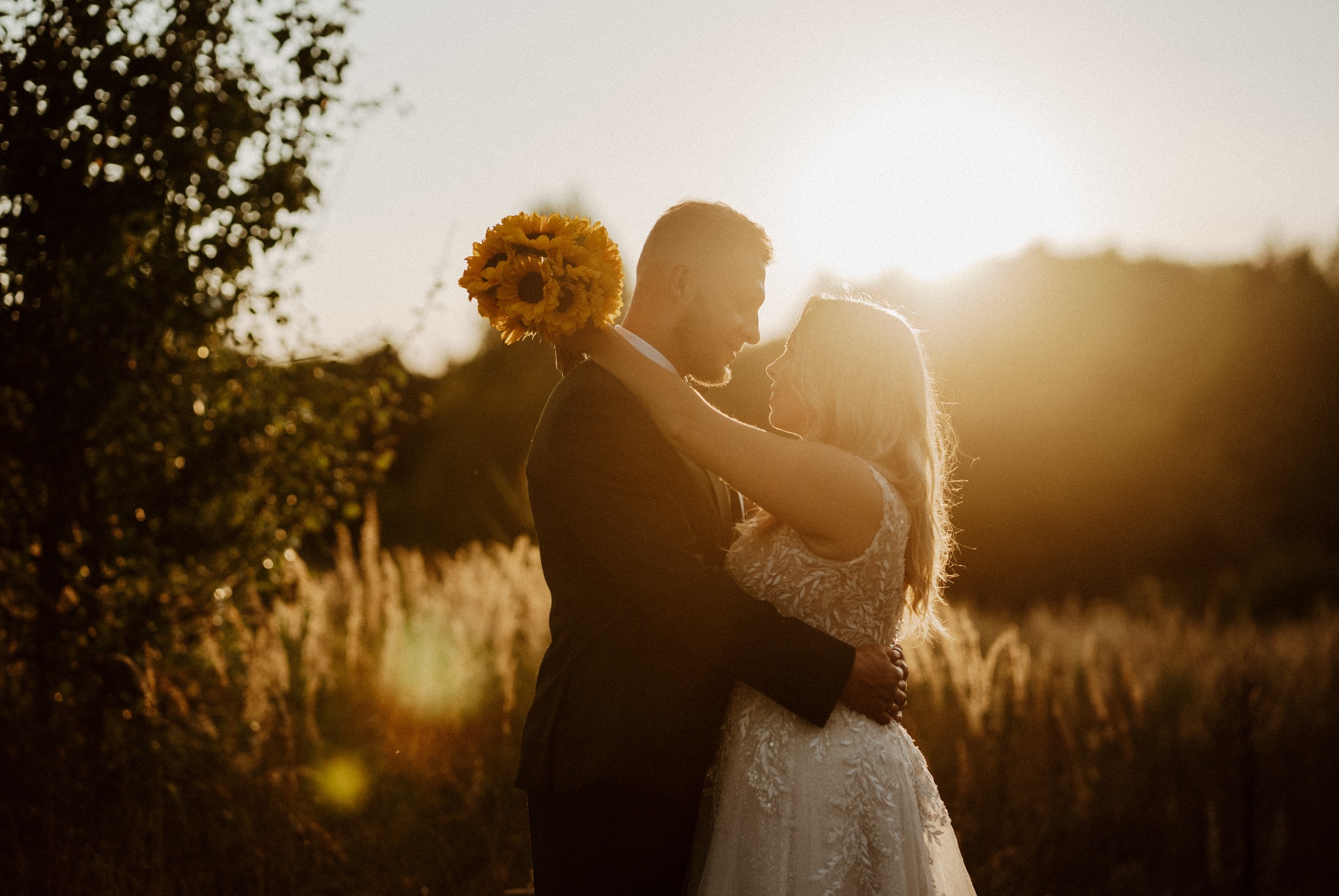 Para młoda w objęciach na tle zachodzącego słońca, panna młoda trzyma bukiet słoneczników, widoczna trawa i drzewa w tle.