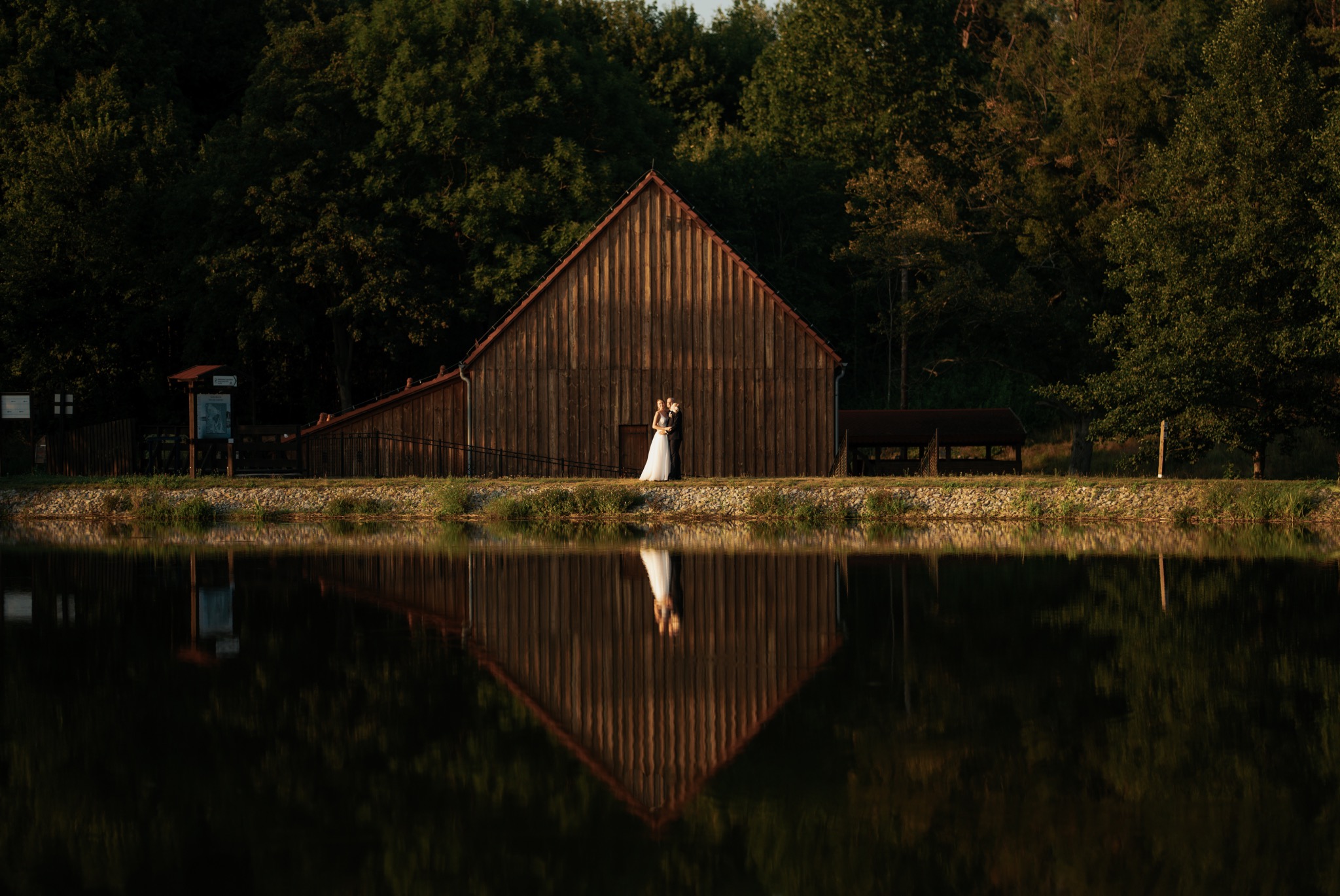 Para młoda w objęciach na tle drewnianego budynku z odbiciem w wodzie, otoczona zielenią drzew