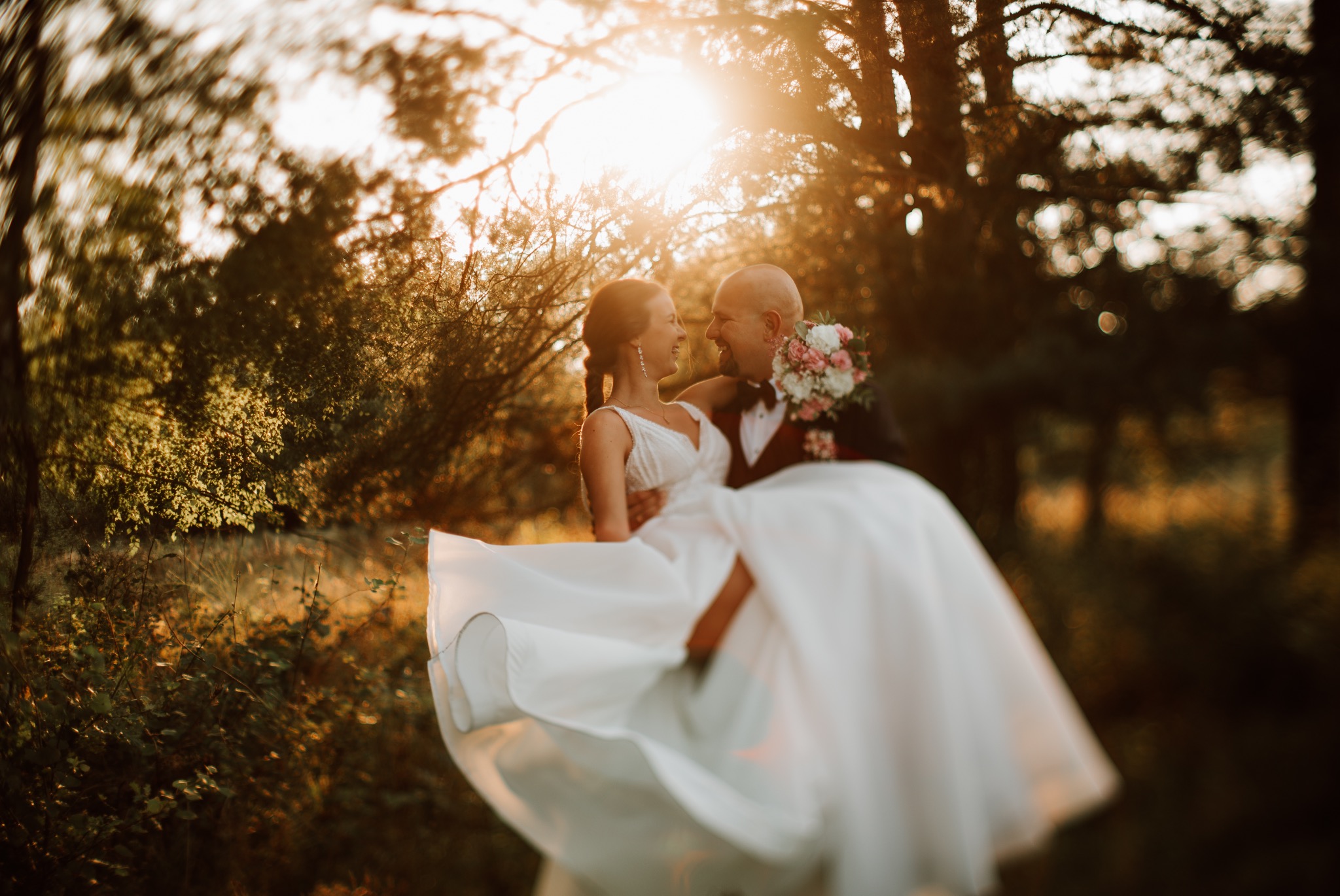 Uśmiechnięta panna młoda w białej sukni jest podnoszona przez pana młodego w burgundowym smokingu na tle zachodzącego słońca w lesie.