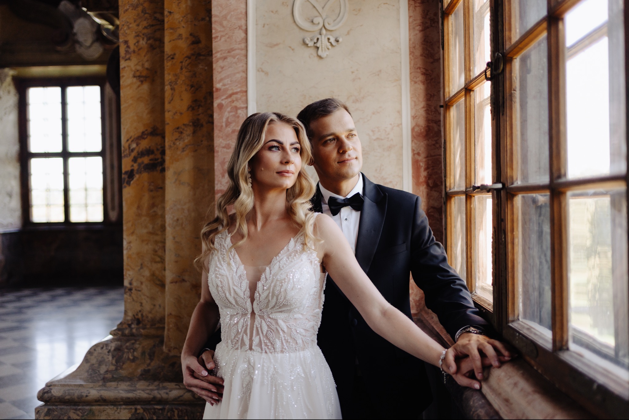 Elegancka sesja narzeczeńska w Strykowie: para młoda w stylowych strojach pozuje przy oknie z kolumnami, w tle marmurowa podłoga i dekoracje.