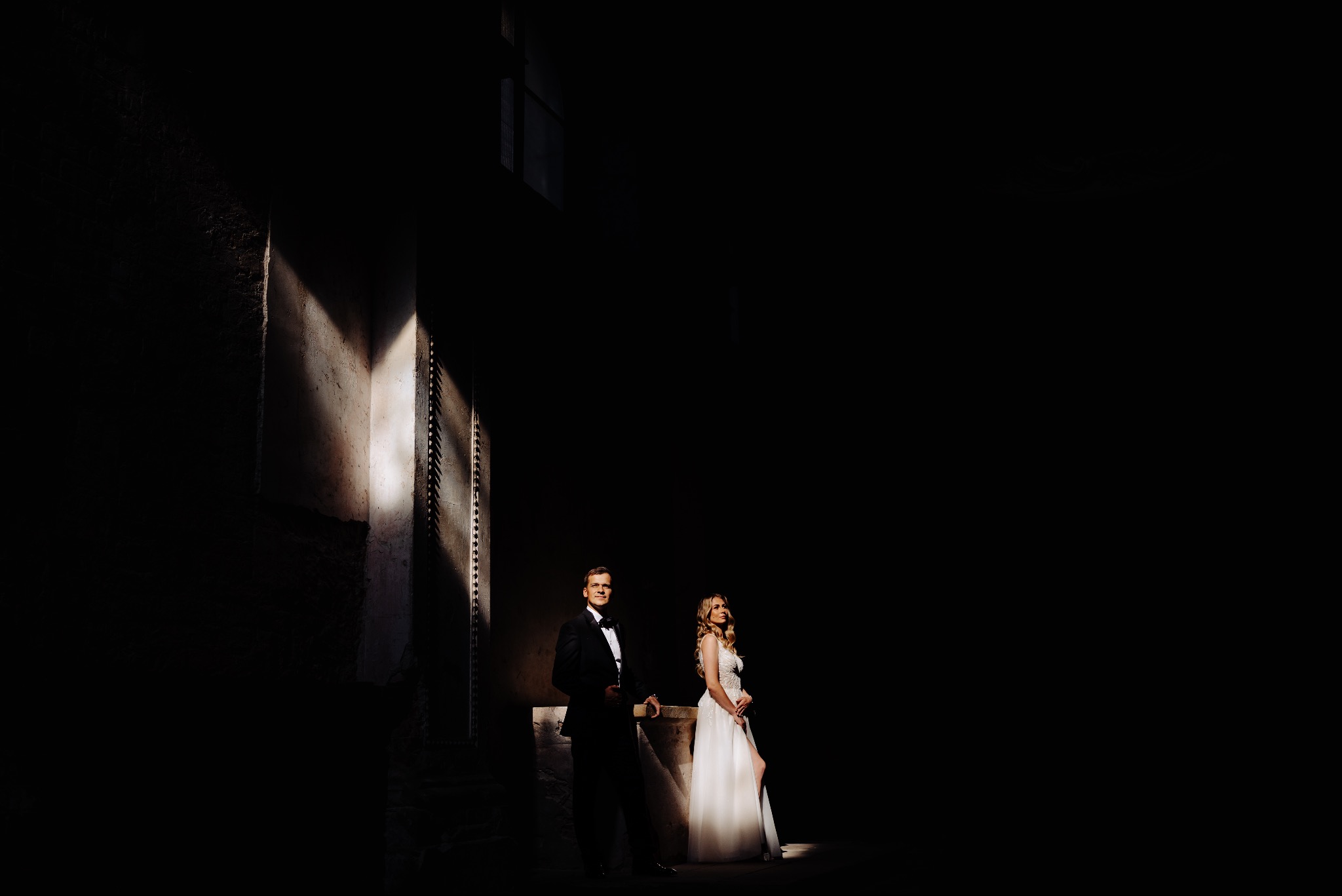 Para młoda w dramatycznym oświetleniu: on w smokingu, ona w białej sukni z rozcięciem, oboje stoją w silnym kontraście światła i cienia na tle surowej architektury.