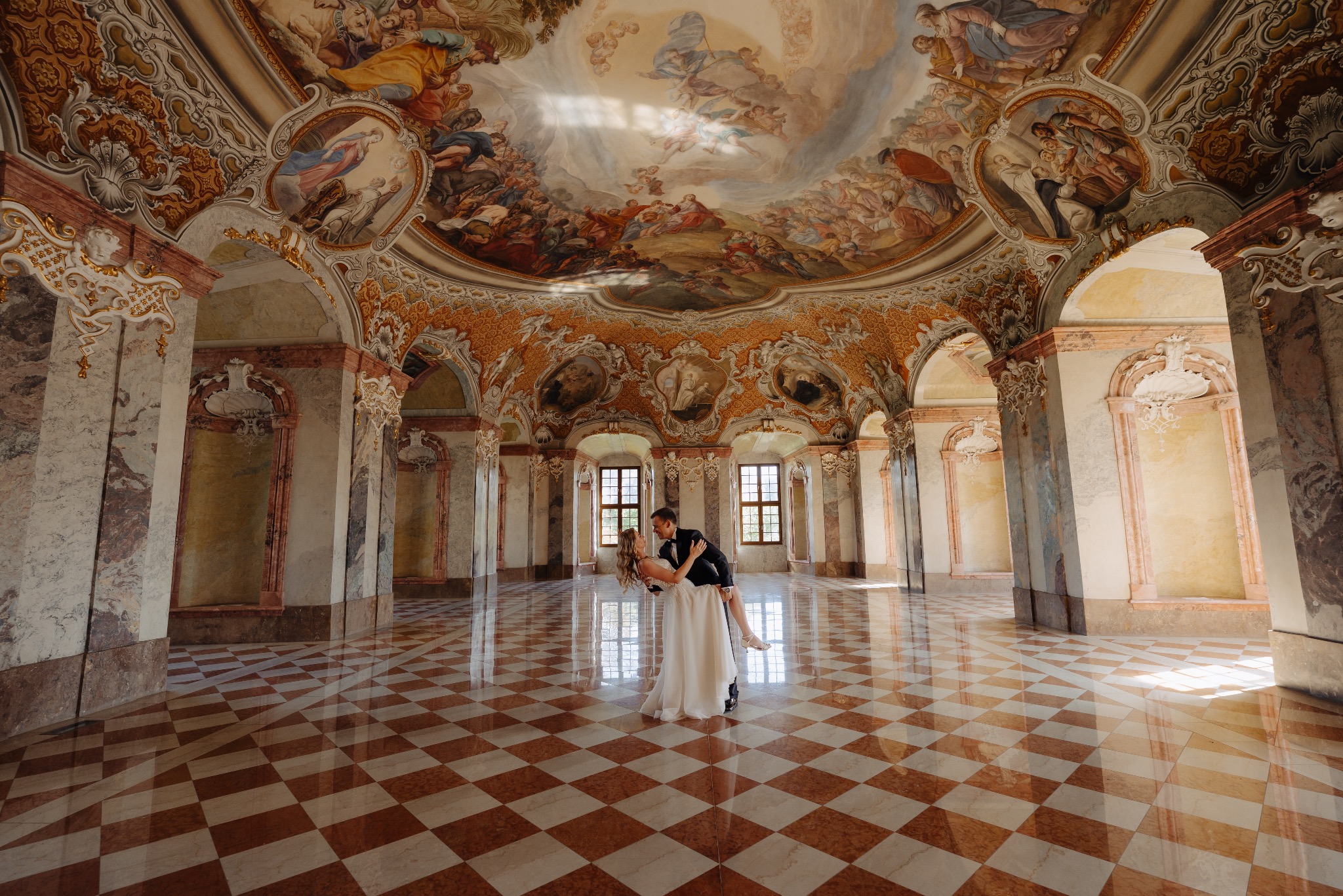 Para młoda w tańcu w barokowym wnętrzu z freskami na suficie i marmurową podłogą w szachownicę. Pan młody trzyma pannę młodą w ramionach.