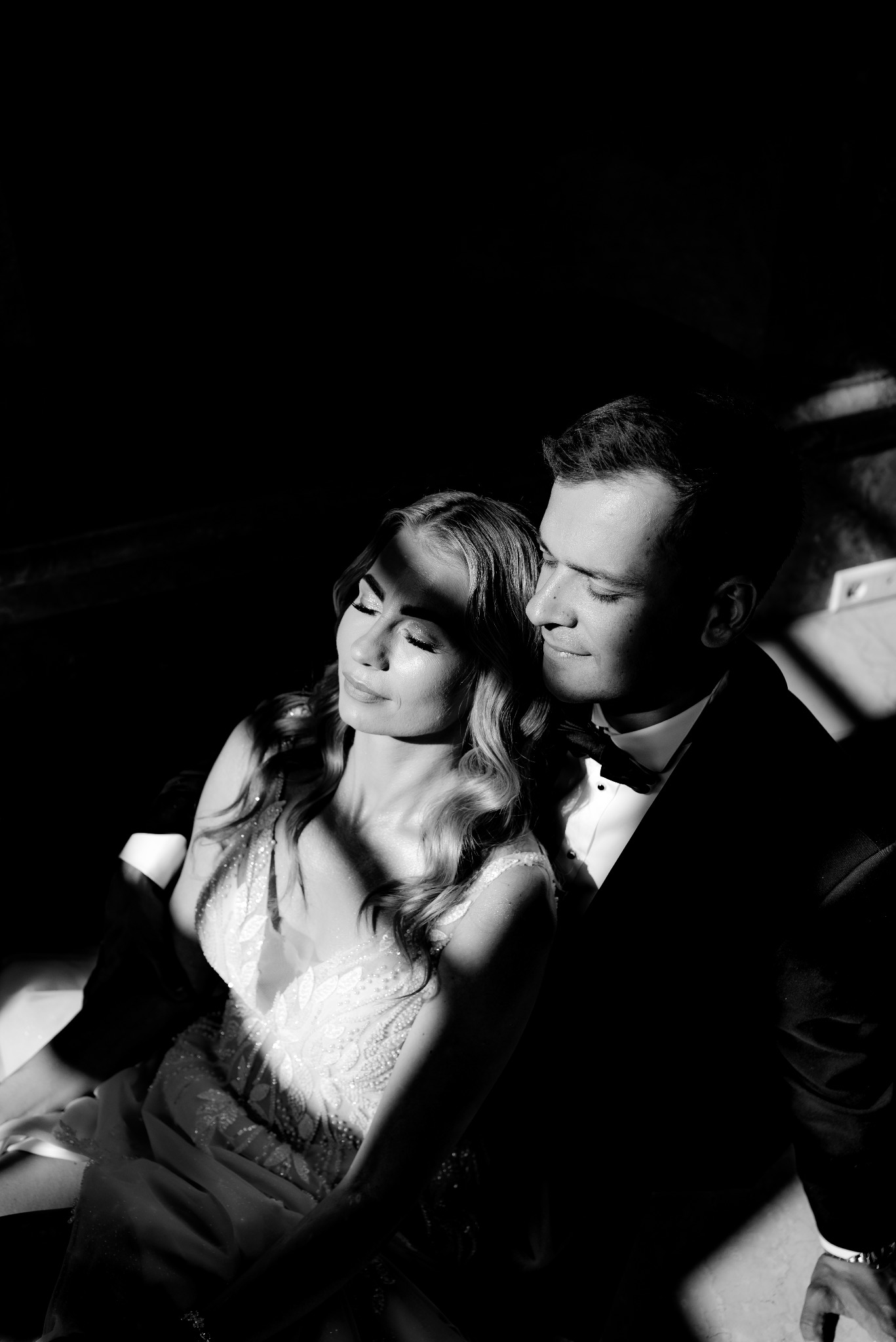 Czarno-białe zdjęcie pary młodej w silnym kontraście światła i cienia. Panna młoda w sukni z koronką, pan młody w smokingu z muchą. Ujęcie z bliska, skupione na twarzach.