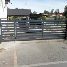 Nowoczesna, przesuwna brama wjazdowa z poziomych profili, szara, widoczna na tle posesji z samochodem i wiatrakiem. Brama zamontowana na posesji z kostki brukowej.