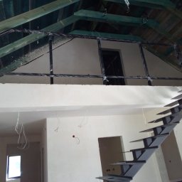 M-Spaw - Wnętrze w trakcie wykańczania z metalowymi schodami o nowoczesnym designie i czarną balustradą na antresoli, zabezpieczoną folią. Widoczne surowe drewno dachu.