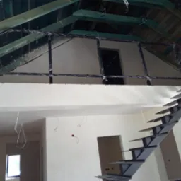 Wnętrze w trakcie wykańczania z metalowymi schodami o nowoczesnym designie i czarną balustradą na antresoli, zabezpieczoną folią. Widoczne surowe drewno dachu.