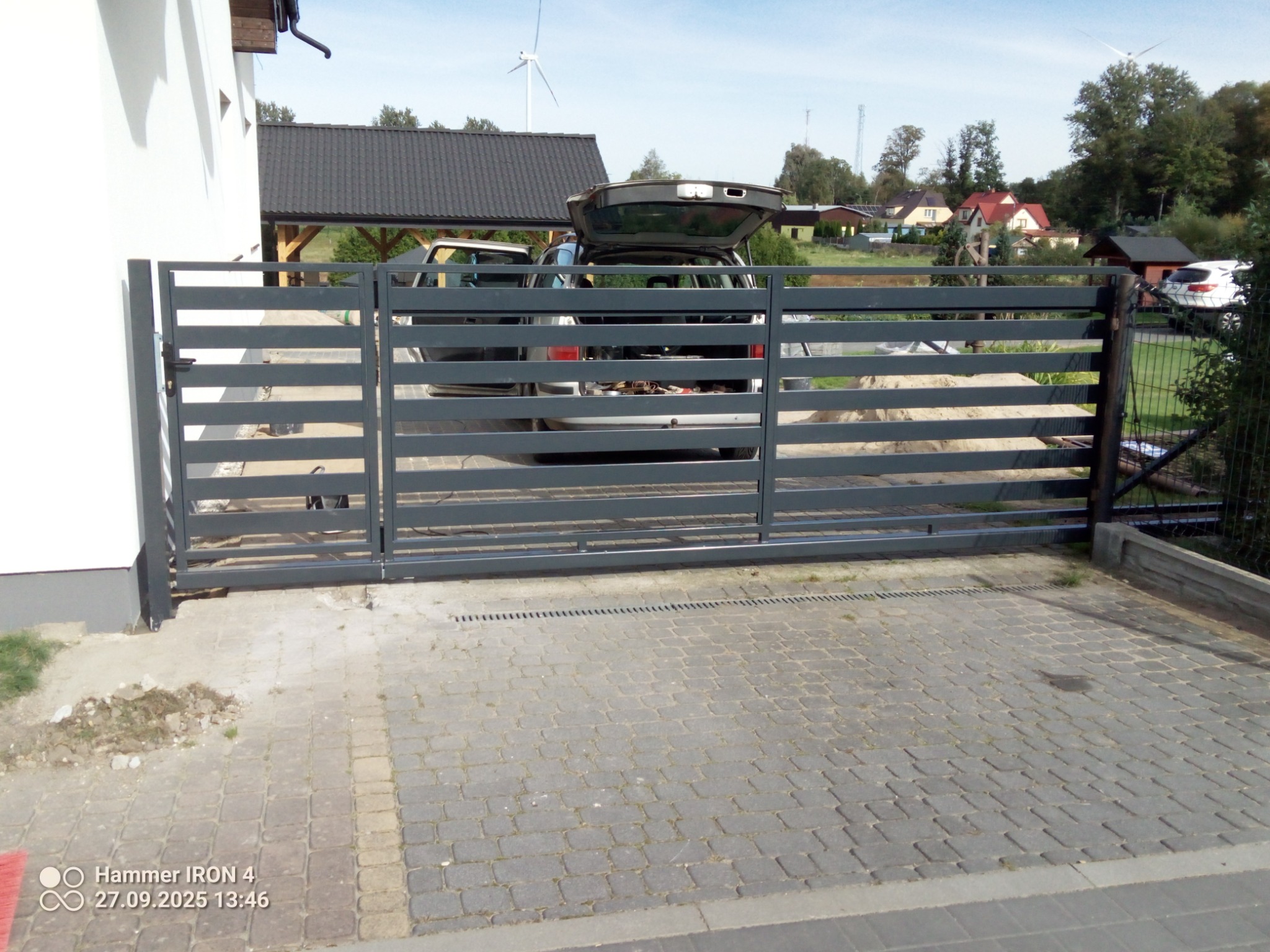 Nowoczesna, przesuwna brama wjazdowa z poziomych profili, szara, widoczna na tle posesji z samochodem i wiatrakiem. Brama zamontowana na posesji z kostki brukowej.