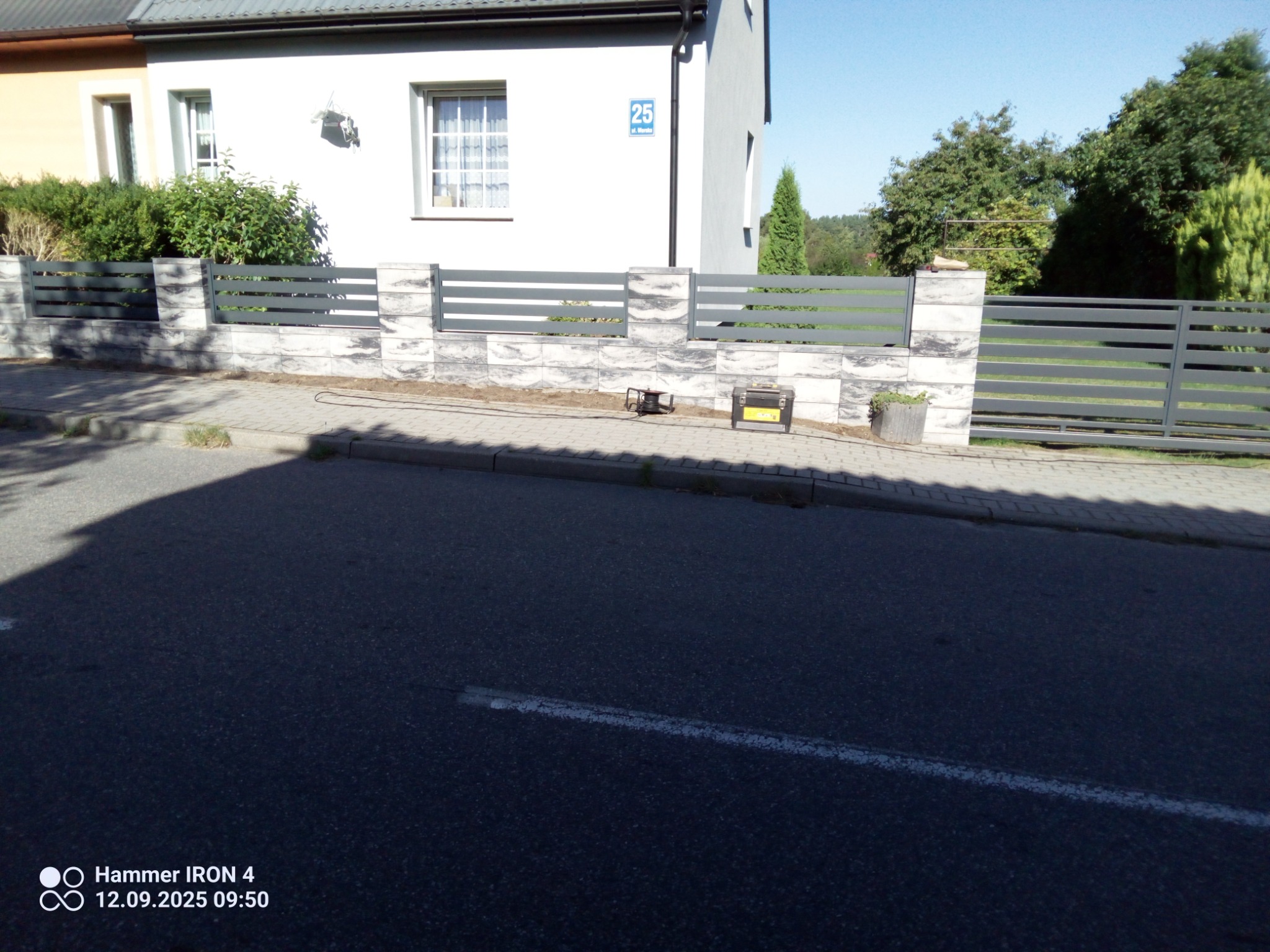 Nowoczesne ogrodzenie panelowe w kolorze grafitowym, z elementami imitującymi kamień, przed domem z numerem 25. Widoczne narzędzia świadczą o świeżym montażu.