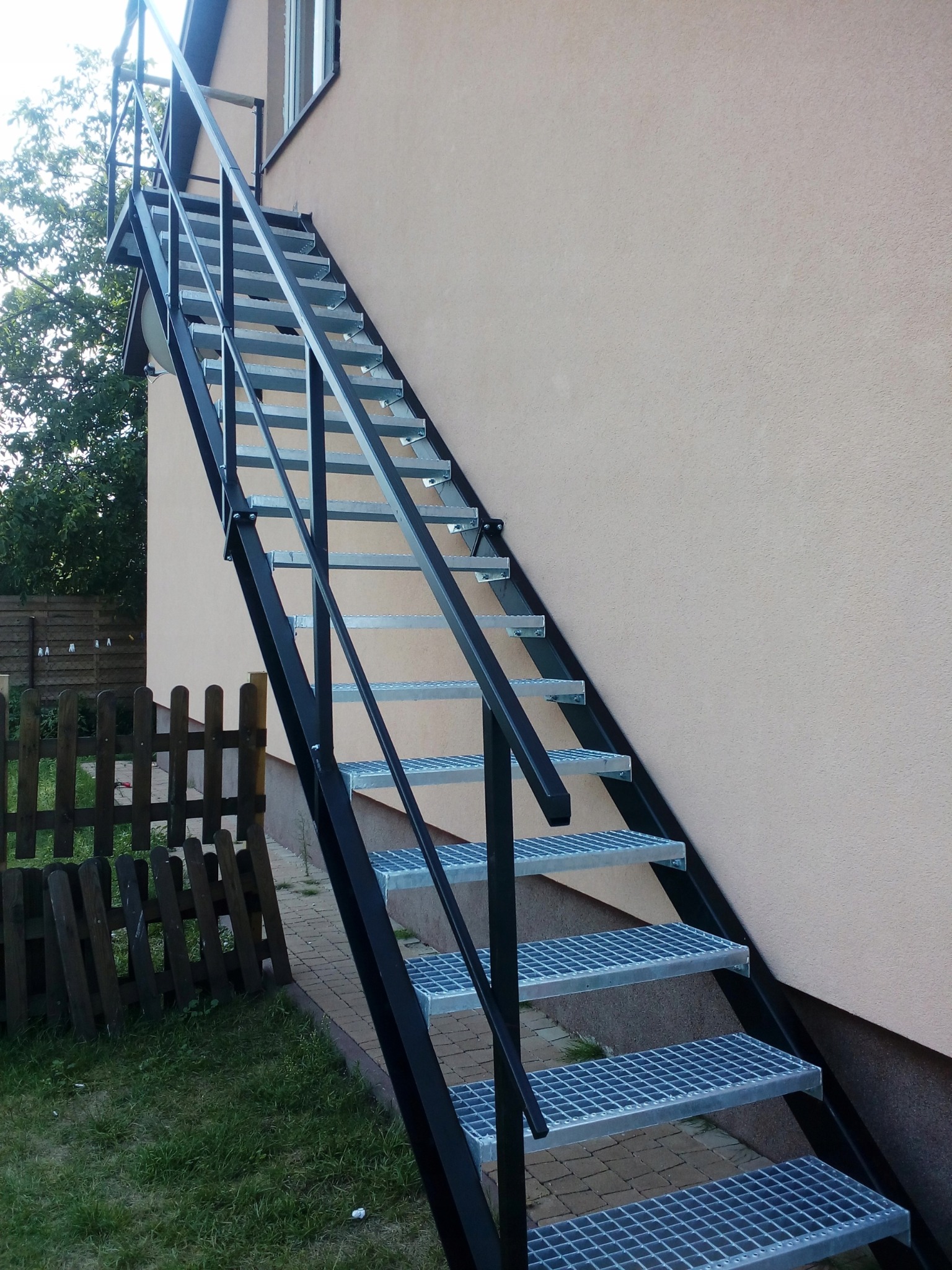 Zewnętrzne schody metalowe z ażurowymi stopniami, pomalowane na czarno, prowadzące do okna na elewacji budynku. Widoczne ogrodzenie i trawnik.