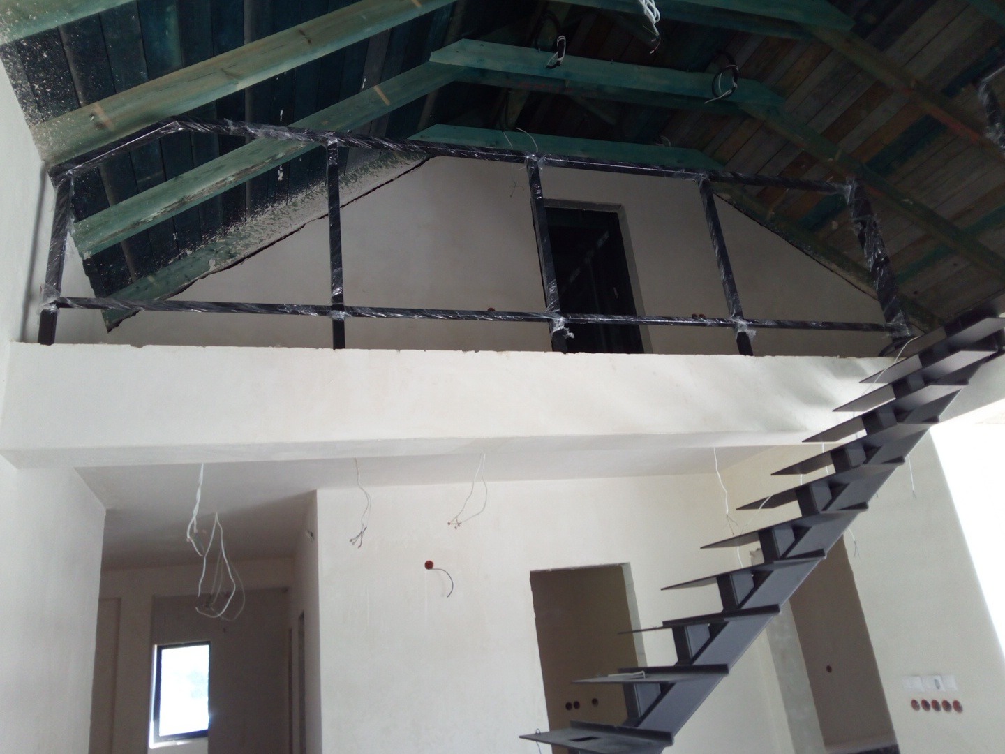 Wnętrze w trakcie wykańczania z metalowymi schodami o nowoczesnym designie i czarną balustradą na antresoli, zabezpieczoną folią. Widoczne surowe drewno dachu.