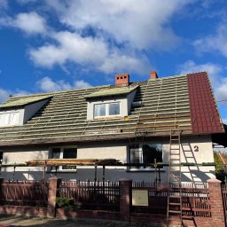 Daniel Komplet Service - Dom jednorodzinny z częściowo zdjętym pokryciem dachu i widoczną konstrukcją drewnianą łat, z fragmentem nowej, bordowej dachówki, rusztowaniem i drabiną opartą o ścianę, na tle błękitnego nieba...